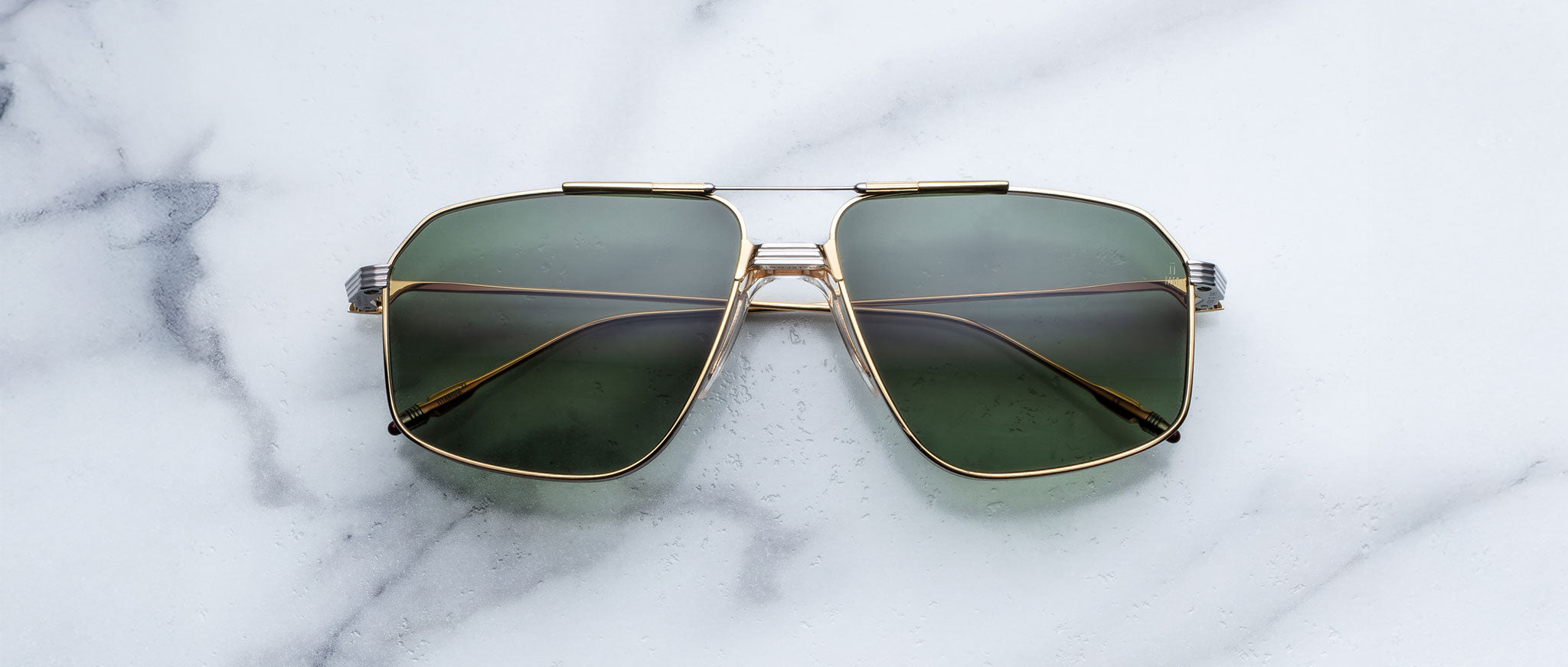 A pair of Jacques Marie Mage Jagger gold-framed, green-tinted oversized aviator sunglasses from Jacques Marie Mage is displayed on a white marble surface.
