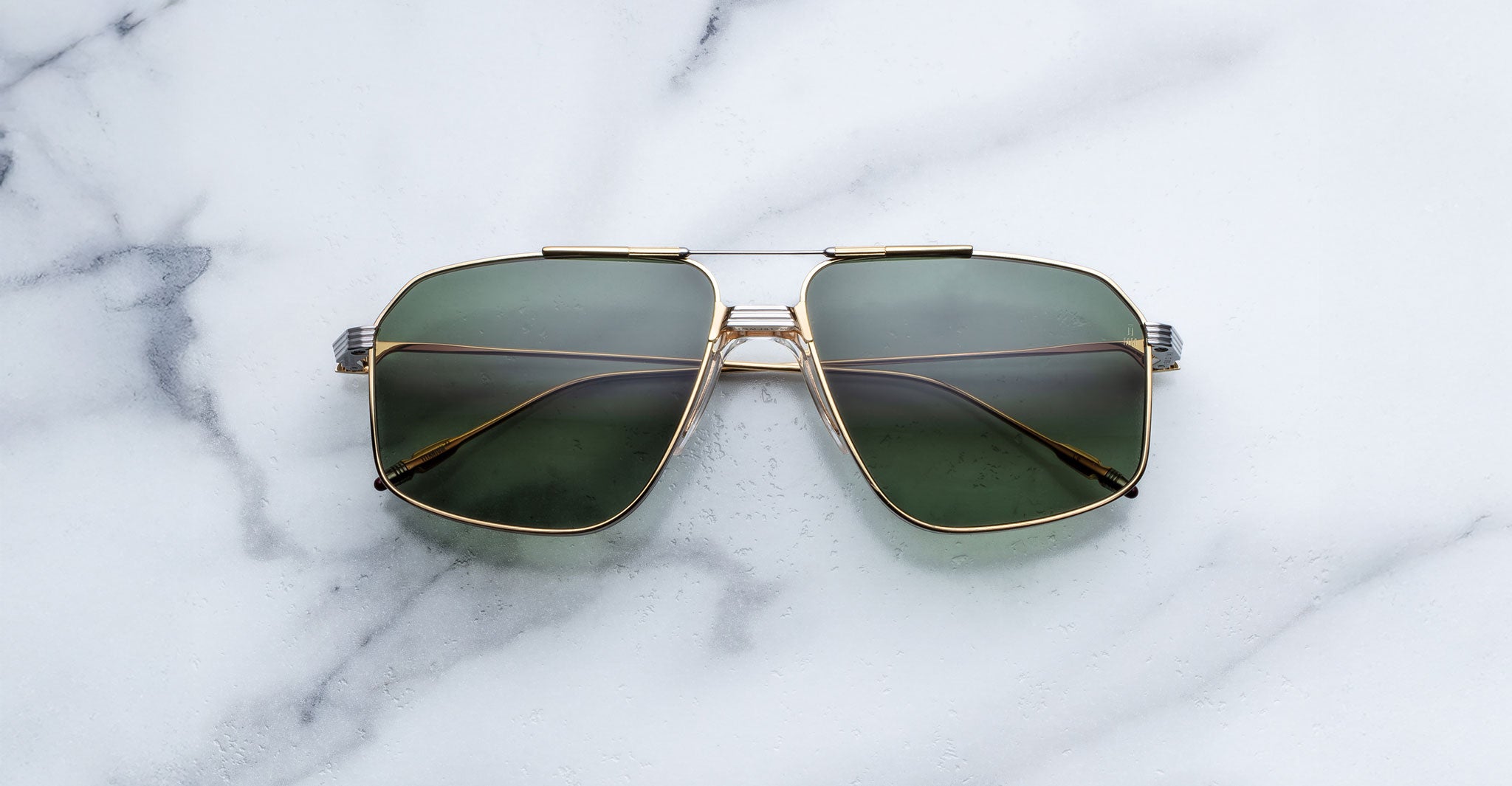 A pair of Jacques Marie Mage Jagger gold-framed, green-tinted oversized aviator sunglasses from Jacques Marie Mage is displayed on a white marble surface.