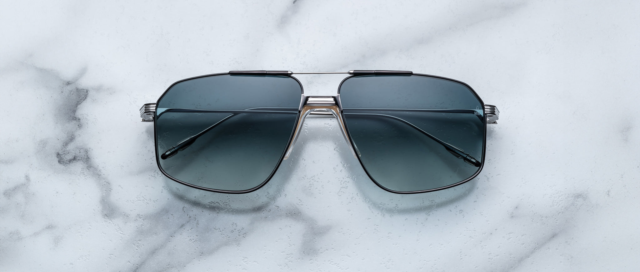 The Jacques Marie Mage Jagger by Jacques Marie Mage features oversized aviator sunglasses with dark tinted lenses and a sleek beta titanium frame, elegantly displayed on a white marble surface.