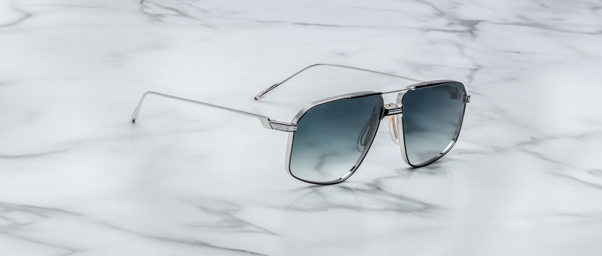 Jacques Marie Mage Jagger aviator sunglasses with silver frames and blue gradient lenses by Jacques Marie Mage rest on a white marble surface.