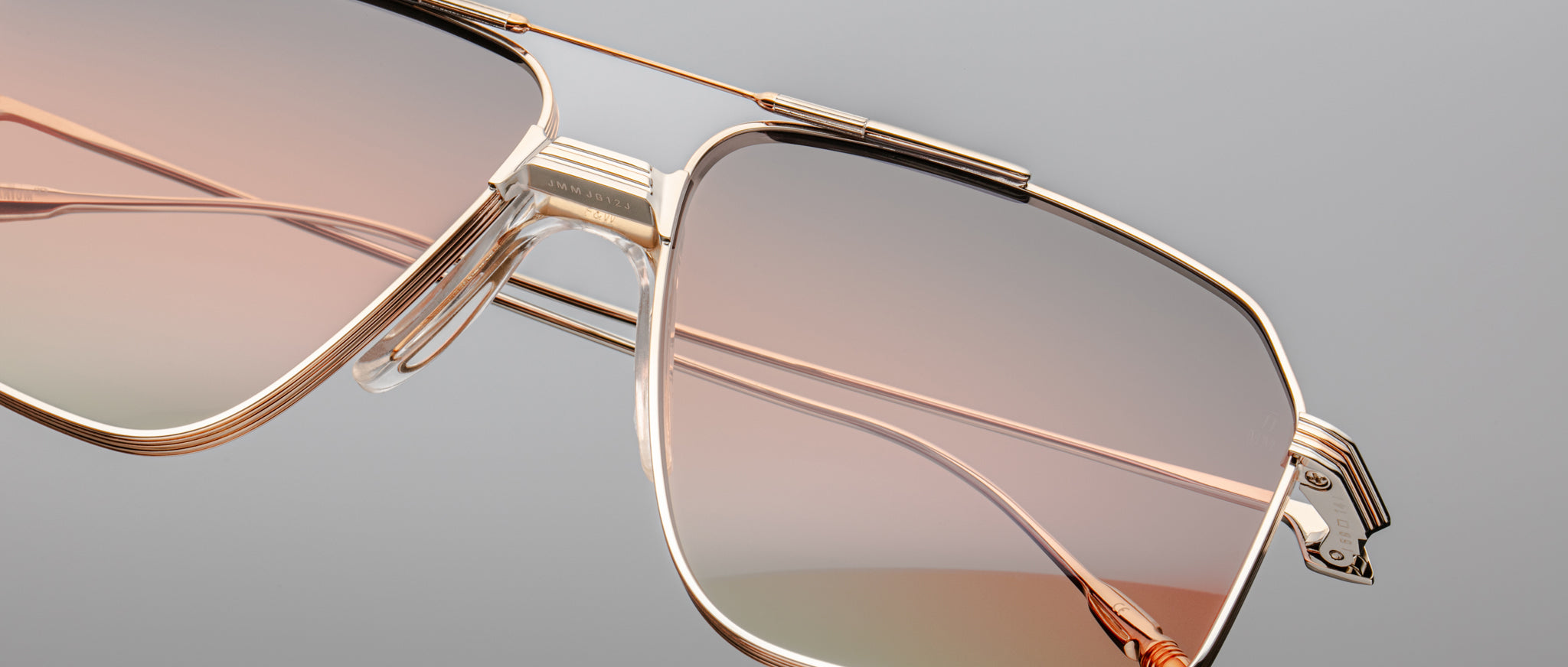Close-up of the Jacques Marie Mage Jagger sunglasses by Jacques Marie Mage, featuring oversized gold beta titanium frames and gradient pink-tinted lenses, shown against a plain gray background.