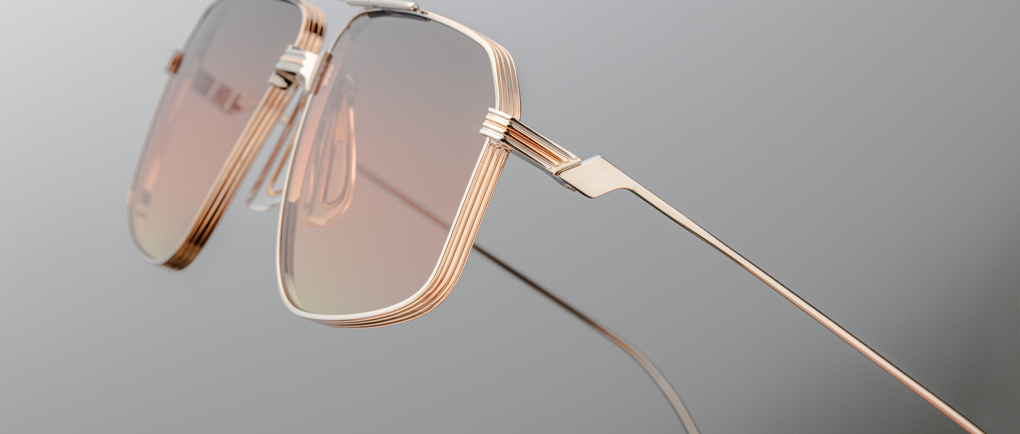 Close-up of Jacques Marie Mage Jagger sunglasses by Jacques Marie Mage, showcasing oversized aviator style with gold metal frames, pink-tinted lenses, and beta titanium construction against a neutral gray background.