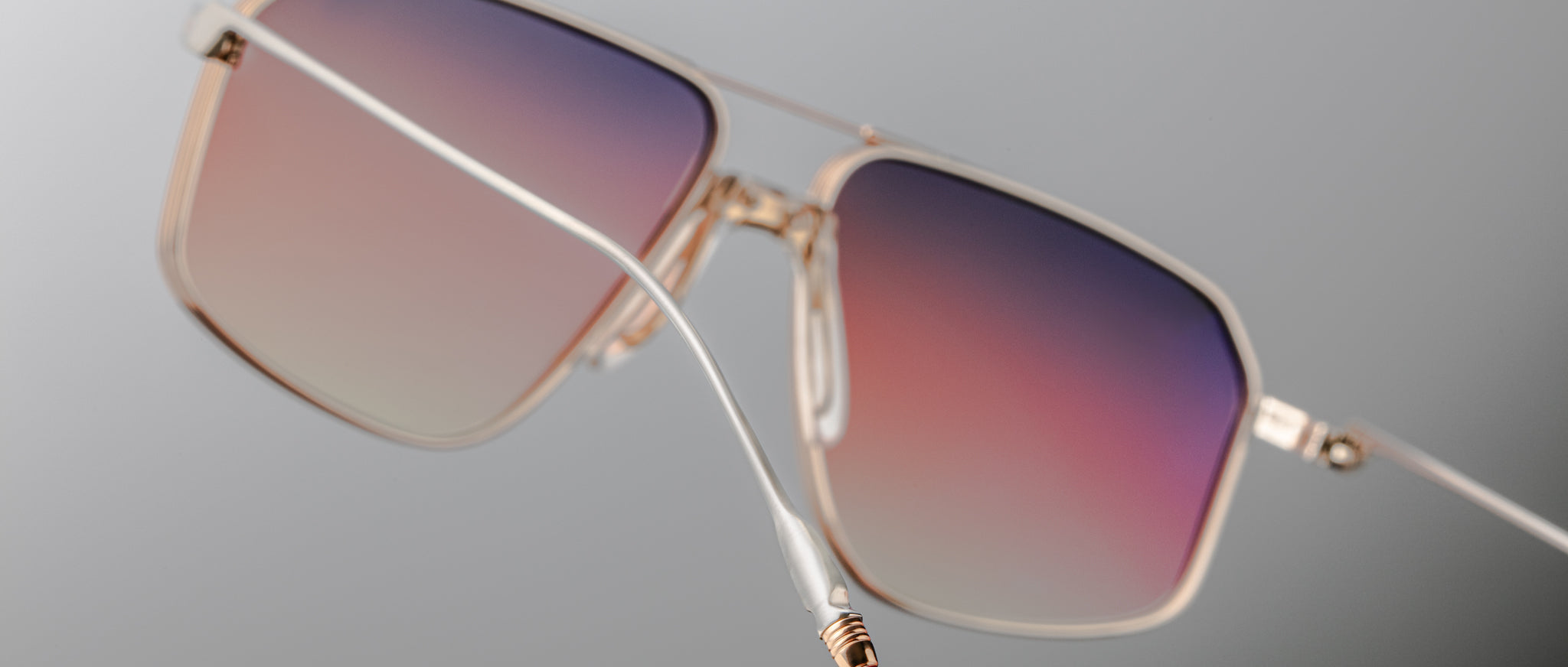 Close-up of Jacques Marie Mage Jagger sunglasses by Jacques Marie Mage, featuring gold rectangular beta titanium frames and gradient lenses fading from purple to pink, set against a neutral background.