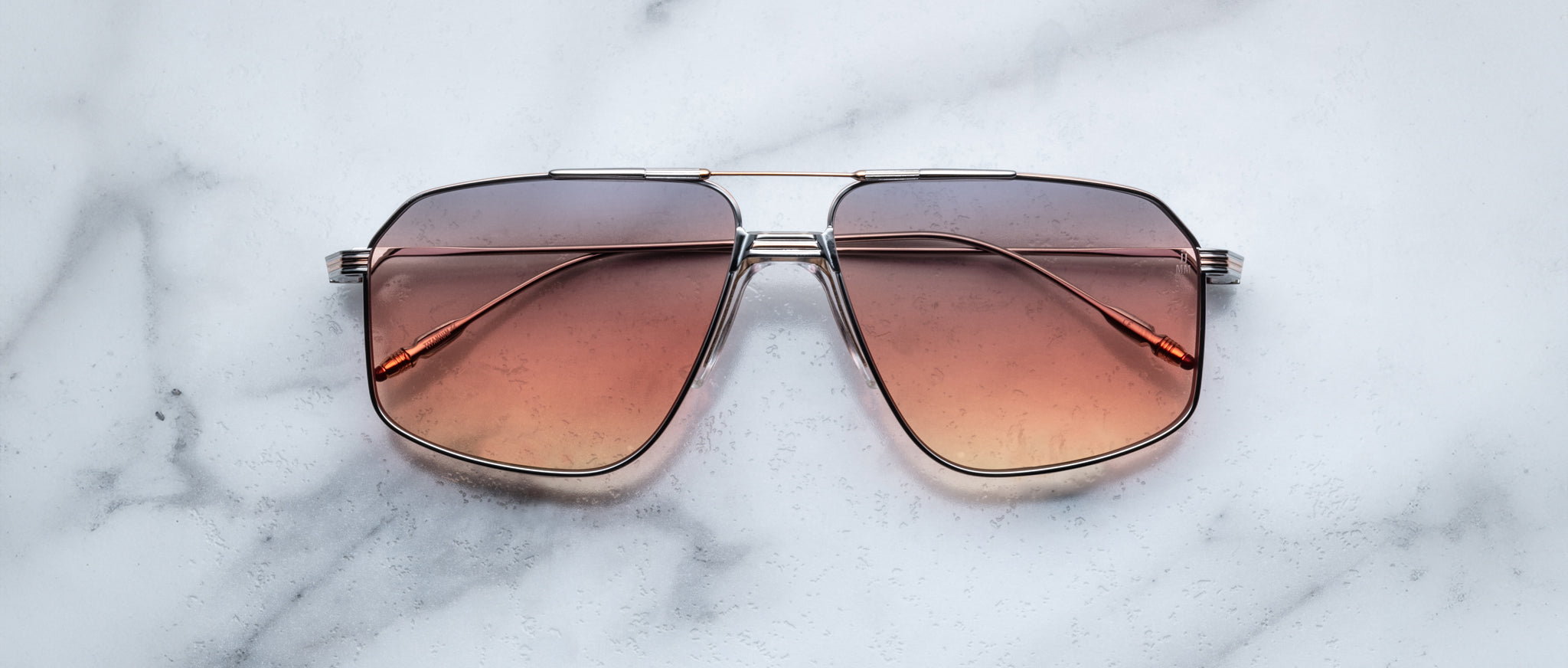 The Jacques Marie Mage Jagger sunglasses by Jacques Marie Mage, featuring oversized aviator gradient brown-tinted lenses and thin beta titanium frames, rest on a white marble surface.