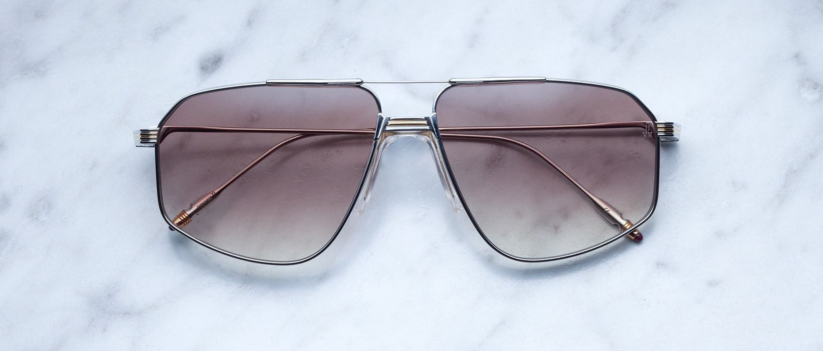A pair of Jacques Marie Mage Jagger oversized aviator sunglasses with brown gradient lenses and thin beta titanium frames from Jacques Marie Mage, displayed on a white marble surface.