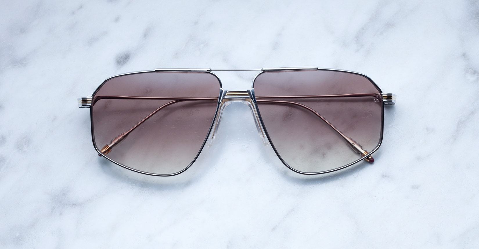 A pair of Jacques Marie Mage Jagger oversized aviator sunglasses with brown gradient lenses and thin beta titanium frames from Jacques Marie Mage, displayed on a white marble surface.
