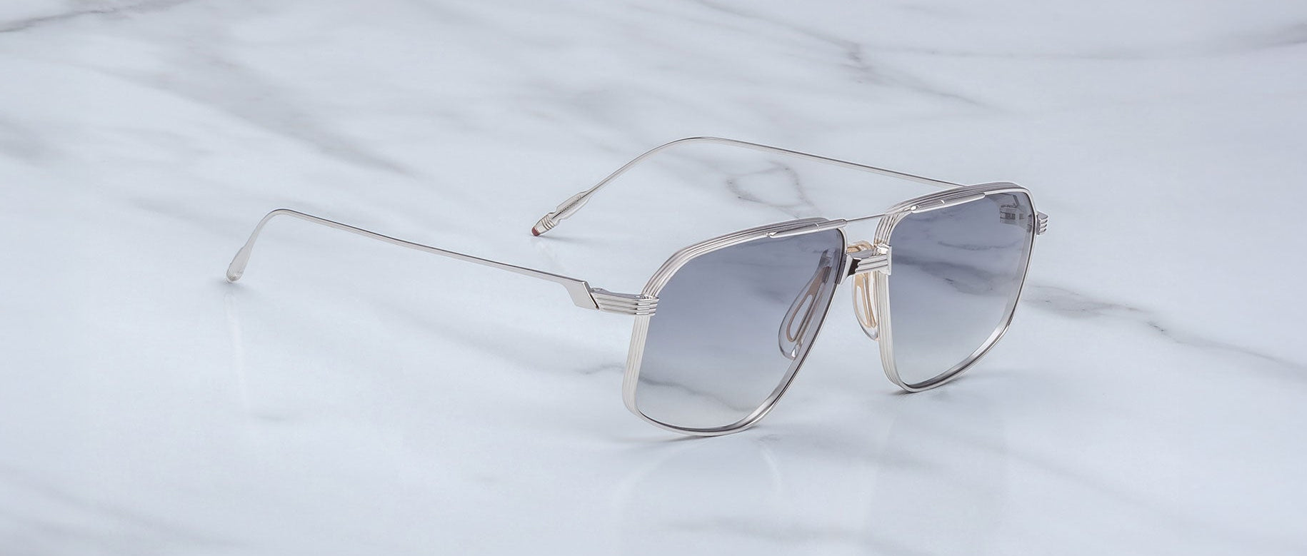 The Jacques Marie Mage Jagger silver metal oversized aviator sunglasses with gray-tinted lenses rest elegantly on a white marble surface.