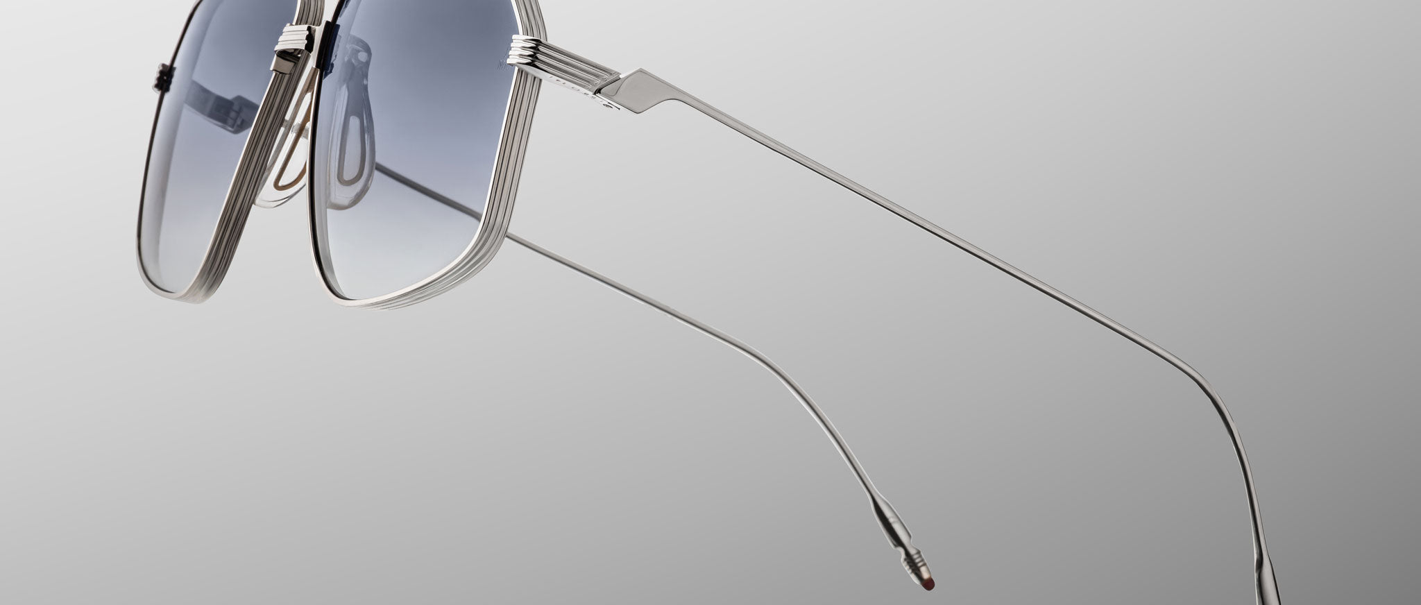 The Jacques Marie Mage Jagger sunglasses by Jacques Marie Mage feature silver oversized aviator frames with blue-tinted lenses, shown against a gray gradient background and angled to showcase thin beta titanium arms.