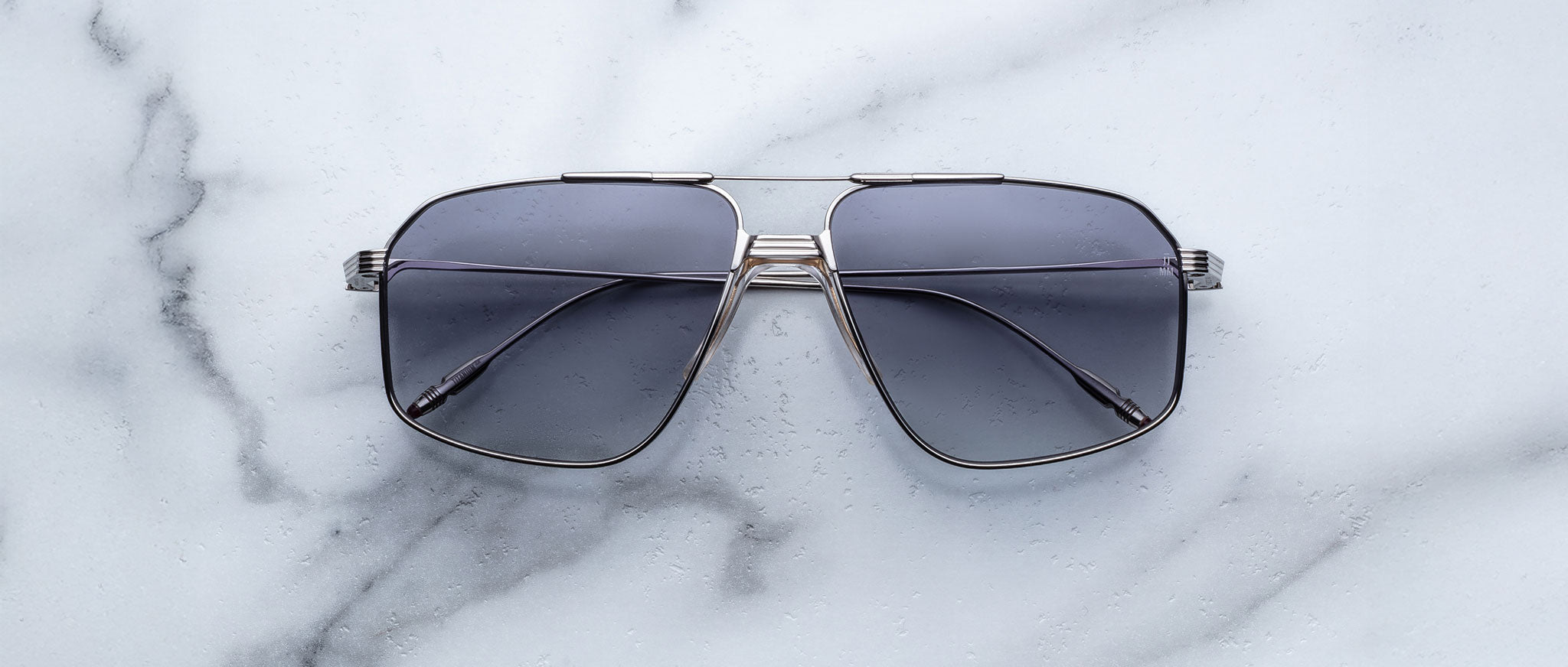 A pair of Jacques Marie Mage Jagger oversized aviator sunglasses with dark lenses and sleek metal frames from Jacques Marie Mage rests on a light marble surface.