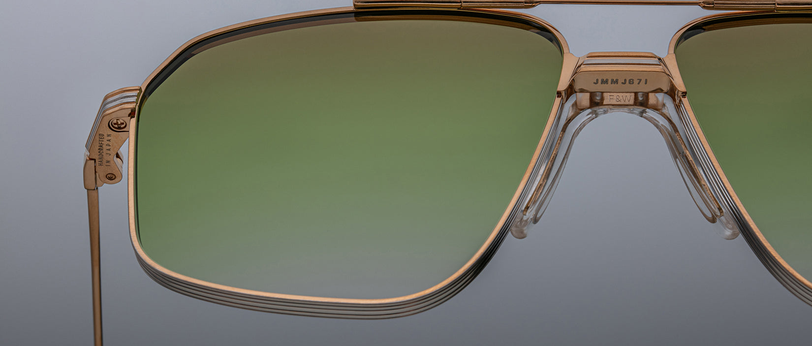 Close-up of Jacques Marie Mage Jagger oversized aviator sunglasses by Jacques Marie Mage, featuring gold frames and green gradient lenses in lightweight beta titanium, set against a neutral gray background.