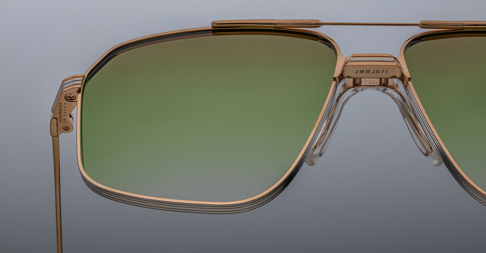 Close-up of Jacques Marie Mage Jagger oversized aviator sunglasses by Jacques Marie Mage, featuring gold frames and green gradient lenses in lightweight beta titanium, set against a neutral gray background.