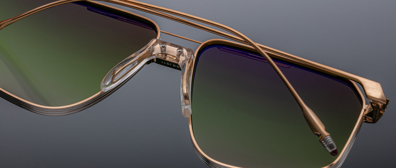 Close-up of Jacques Marie Mage Jagger oversized aviator sunglasses by Jacques Marie Mage, with gold beta titanium frames and green-to-purple gradient lenses, highlighting frame and nose pads against a dark background.