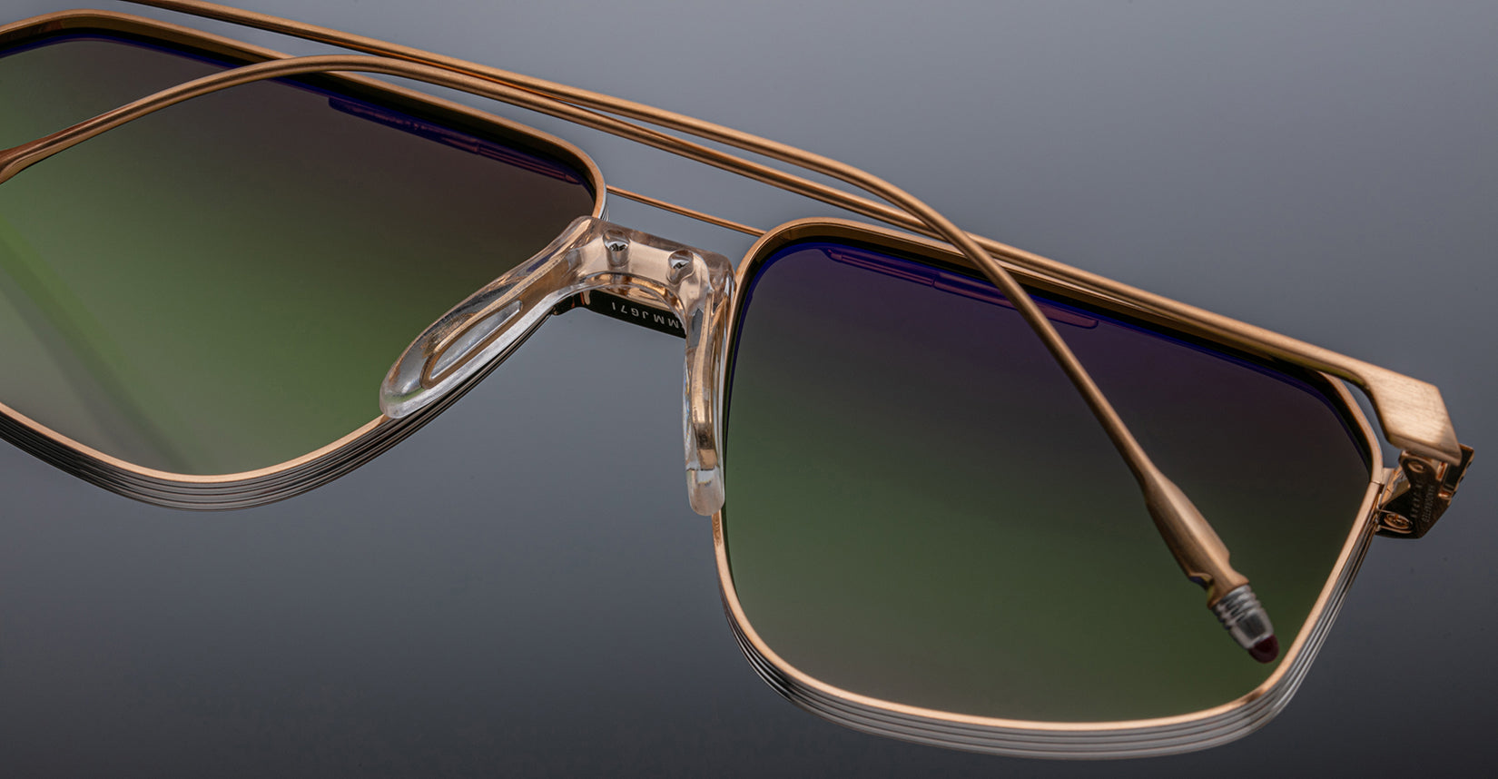 Close-up of Jacques Marie Mage Jagger oversized aviator sunglasses by Jacques Marie Mage, with gold beta titanium frames and green-to-purple gradient lenses, highlighting frame and nose pads against a dark background.