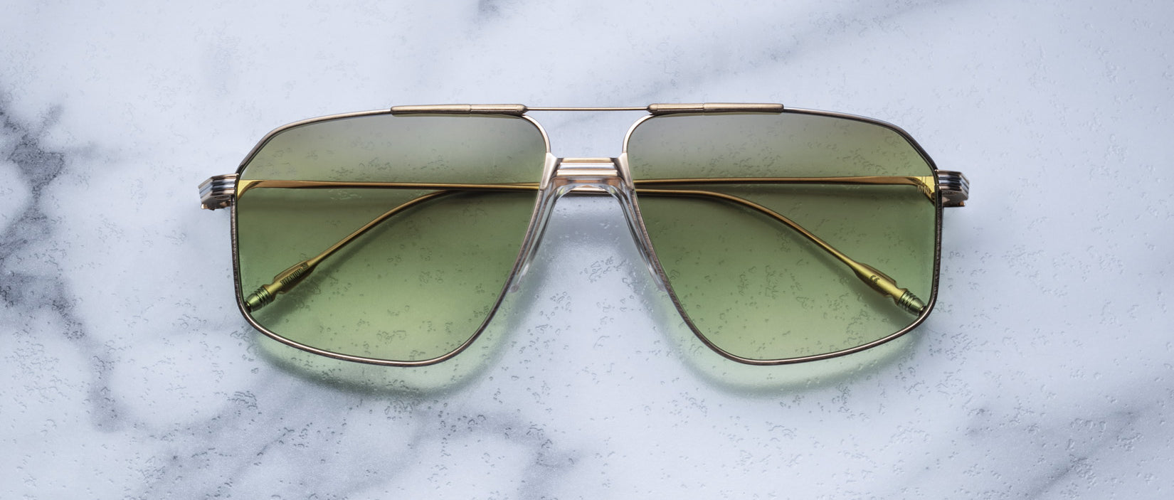 The Jacques Marie Mage Jagger sunglasses by Jacques Marie Mage, featuring gold frames and green-tinted oversized aviator lenses, rest elegantly on a white marble surface.