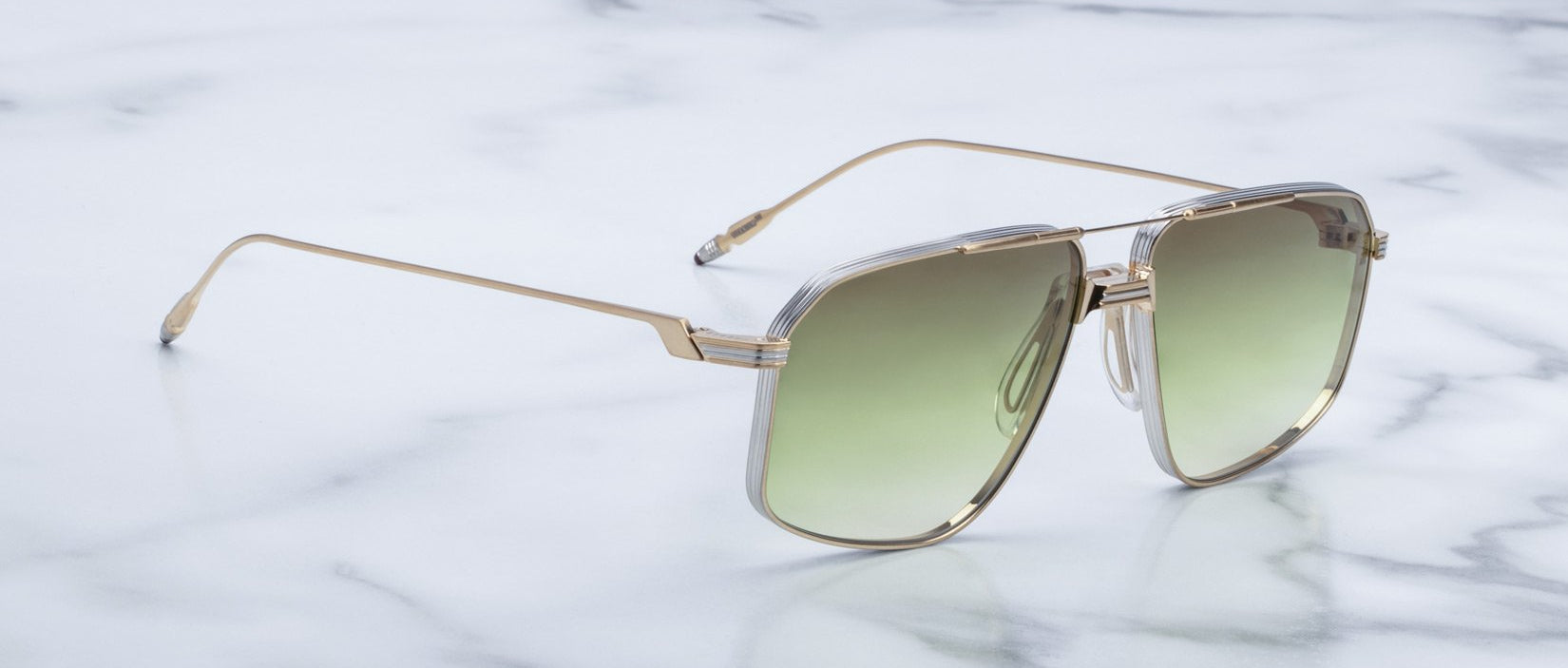 A pair of Jacques Marie Mage Jagger sunglasses by Jacques Marie Mage, featuring oversized gold metal aviator frames and green gradient lenses, is displayed on a white marble surface.