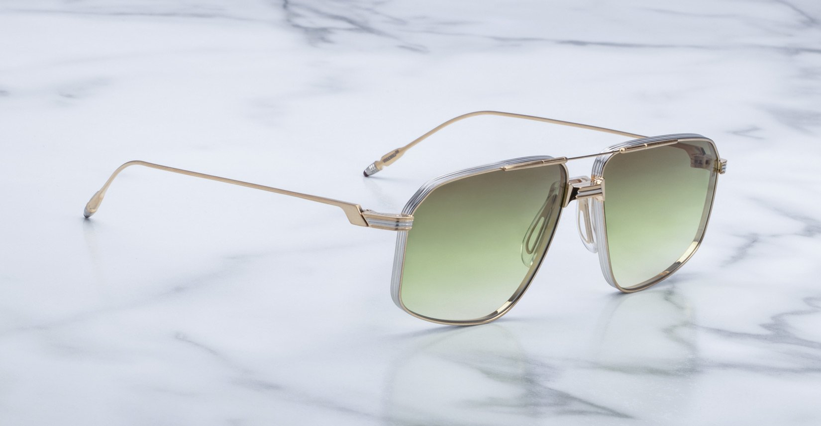 A pair of Jacques Marie Mage Jagger sunglasses by Jacques Marie Mage, featuring oversized gold metal aviator frames and green gradient lenses, is displayed on a white marble surface.
