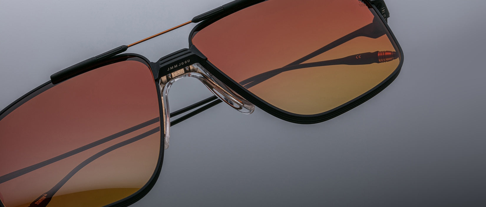 Close-up of Jacques Marie Mage Jagger sunglasses: oversized aviator style with black frames, gradient orange-brown lenses, and beta titanium accents on the bridge, nose pads, and temples, set against a grey background.