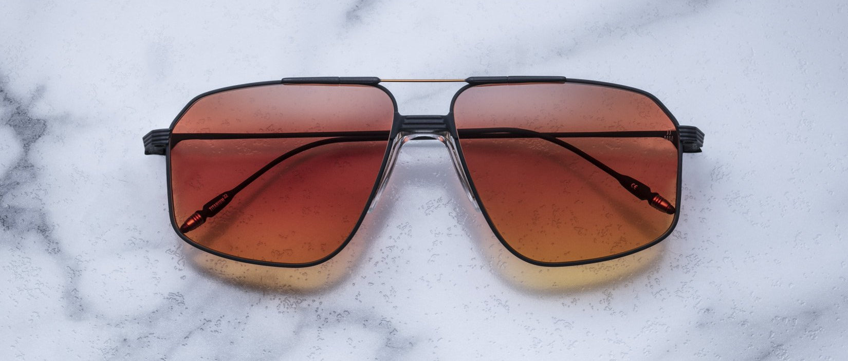 The Jacques Marie Mage Jagger by Jacques Marie Mage are oversized aviator sunglasses with gradient orange-brown lenses, a black metal beta titanium frame, shown on a white marble surface.