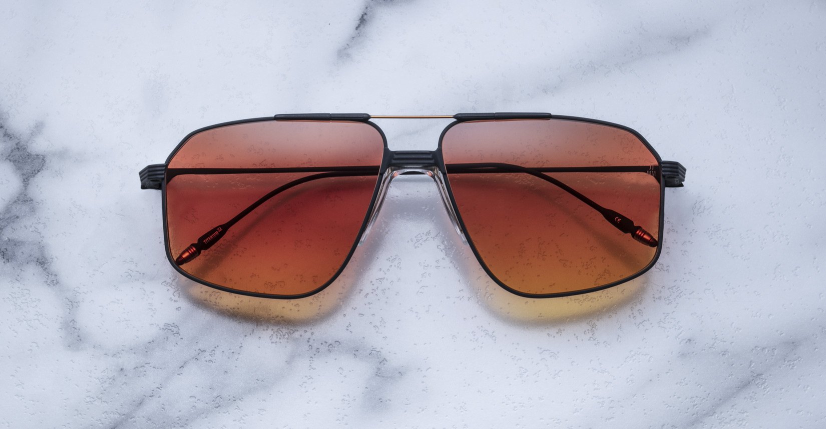 The Jacques Marie Mage Jagger by Jacques Marie Mage are oversized aviator sunglasses with gradient orange-brown lenses, a black metal beta titanium frame, shown on a white marble surface.