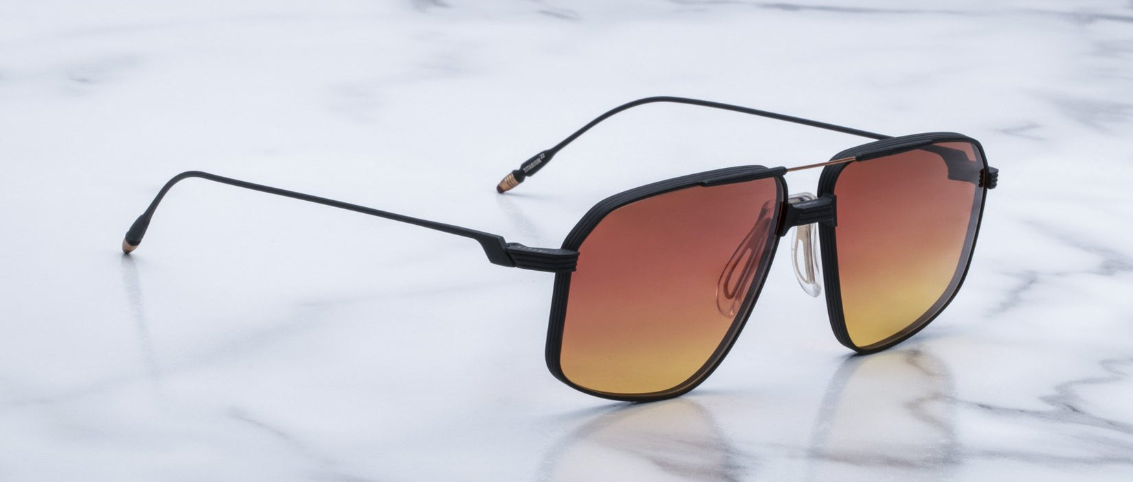 The Jacques Marie Mage Jagger sunglasses feature square black frames and gradient orange lenses, inspired by the signature style of Jacques Marie Mage, and are elegantly displayed on a white marble surface.