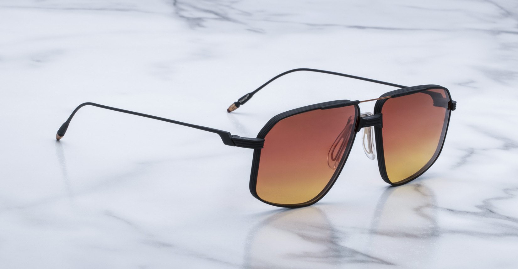 The Jacques Marie Mage Jagger sunglasses feature square black frames and gradient orange lenses, inspired by the signature style of Jacques Marie Mage, and are elegantly displayed on a white marble surface.