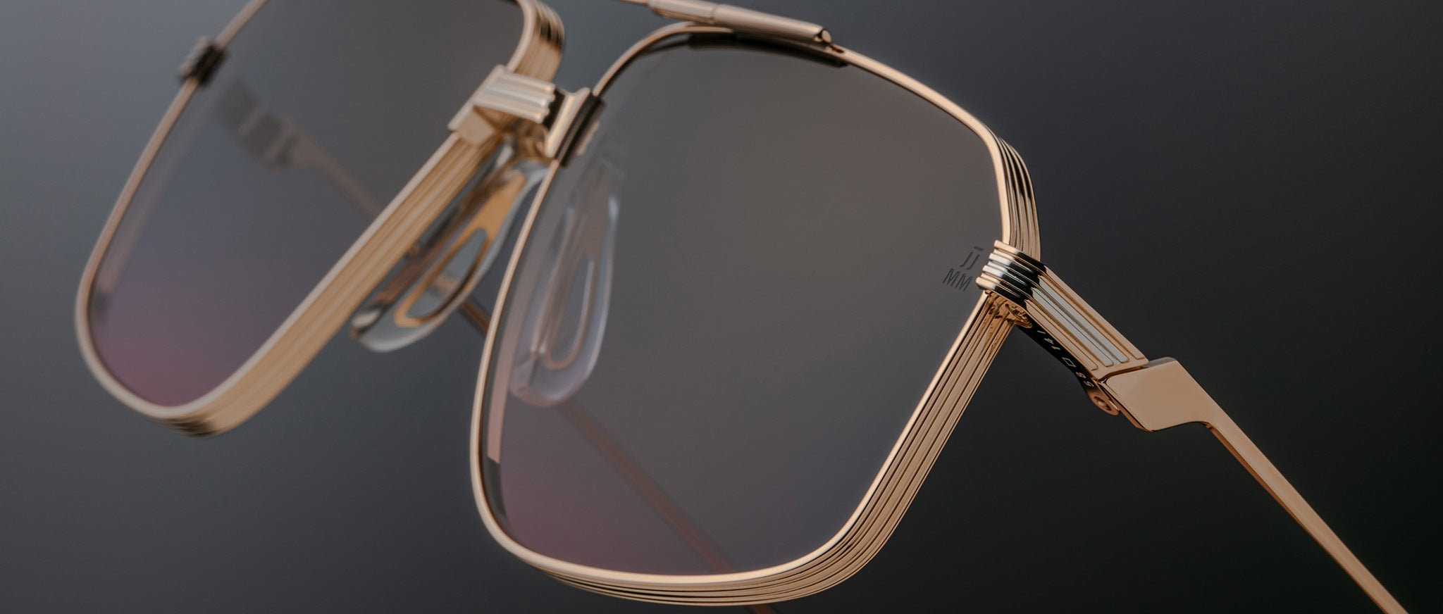 Close-up of Jacques Marie Mage Jagger aviator sunglasses by Jacques Marie Mage, featuring a gold geometric beta titanium frame, tinted lenses, and prominent nose pads, inspired by the iconic Jagger style against a dark background.