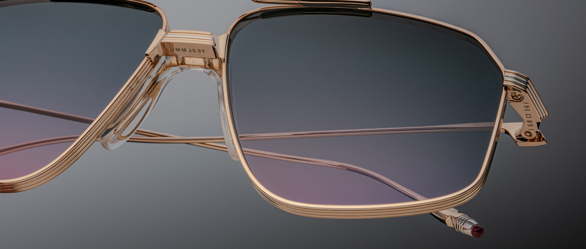 Close-up of oversized Jacques Marie Mage Jagger aviator sunglasses, featuring gold frames and lightweight gradient blue-to-purple tinted beta titanium lenses against a neutral background.