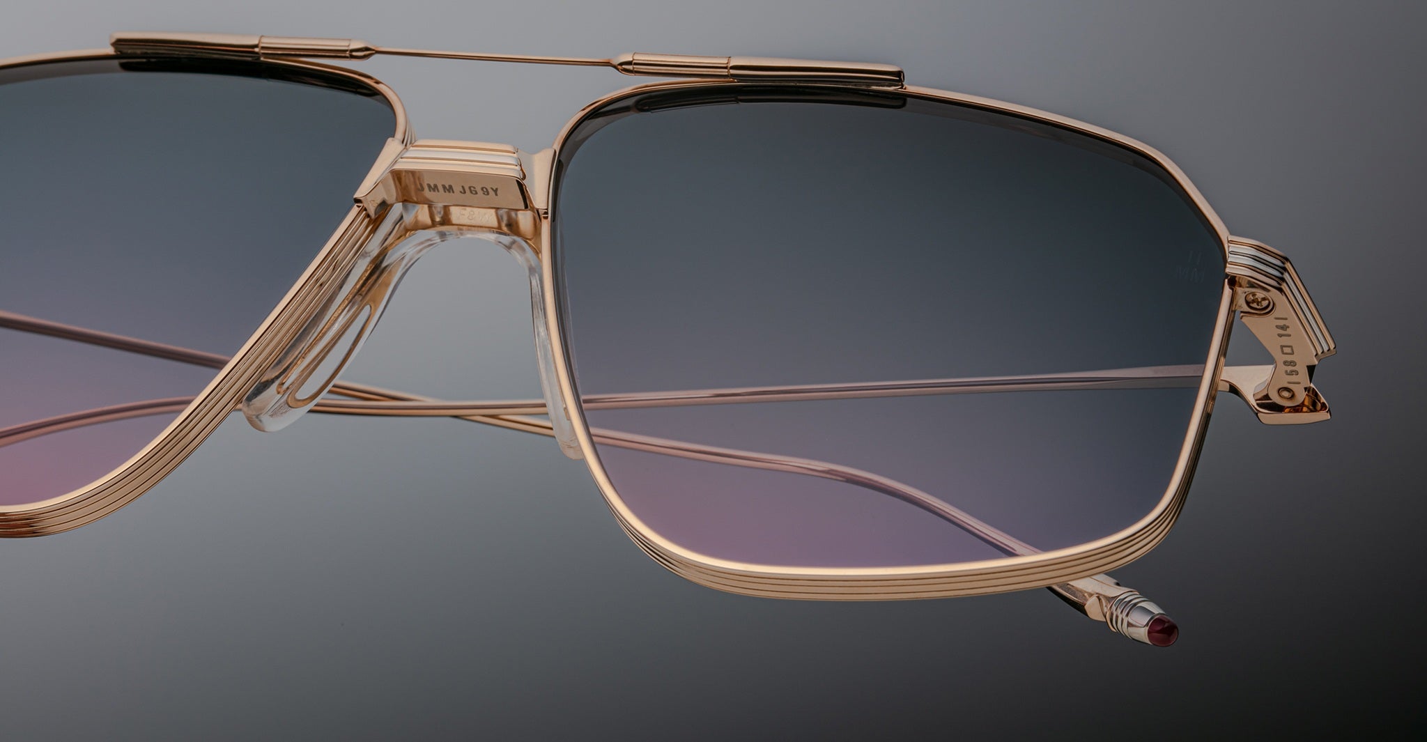 Close-up of oversized Jacques Marie Mage Jagger aviator sunglasses, featuring gold frames and lightweight gradient blue-to-purple tinted beta titanium lenses against a neutral background.