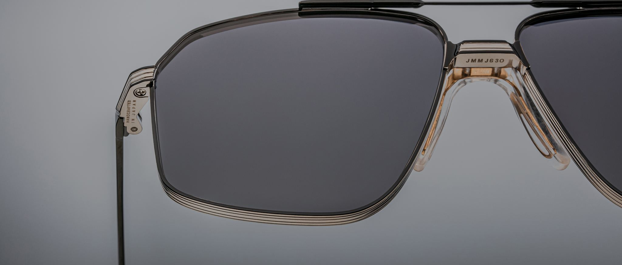 Close-up of Jacques Marie Mage Jagger sunglasses by Jacques Marie Mage, featuring oversized aviator style, dark lenses, a beta titanium metal frame, and visible hinge detail on a neutral gray background.
