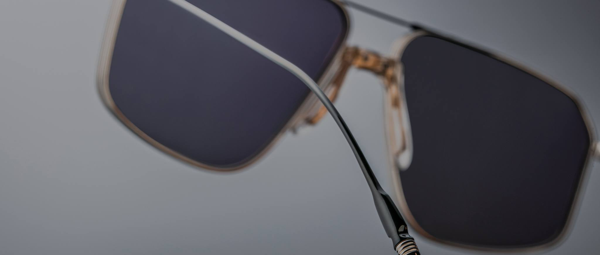 Close-up of Jacques Marie Mage Jagger sunglasses by Jacques Marie Mage, featuring black rectangular frames, gold accents, beta titanium details, and a red temple tip against a blurred gray background.