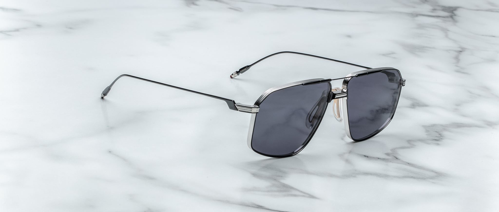A pair of Jacques Marie Mage Jagger oversized aviator sunglasses with dark lenses and beta titanium accents rests stylishly on a white marble surface, reflecting the bold elegance of the Jacques Marie Mage brand.