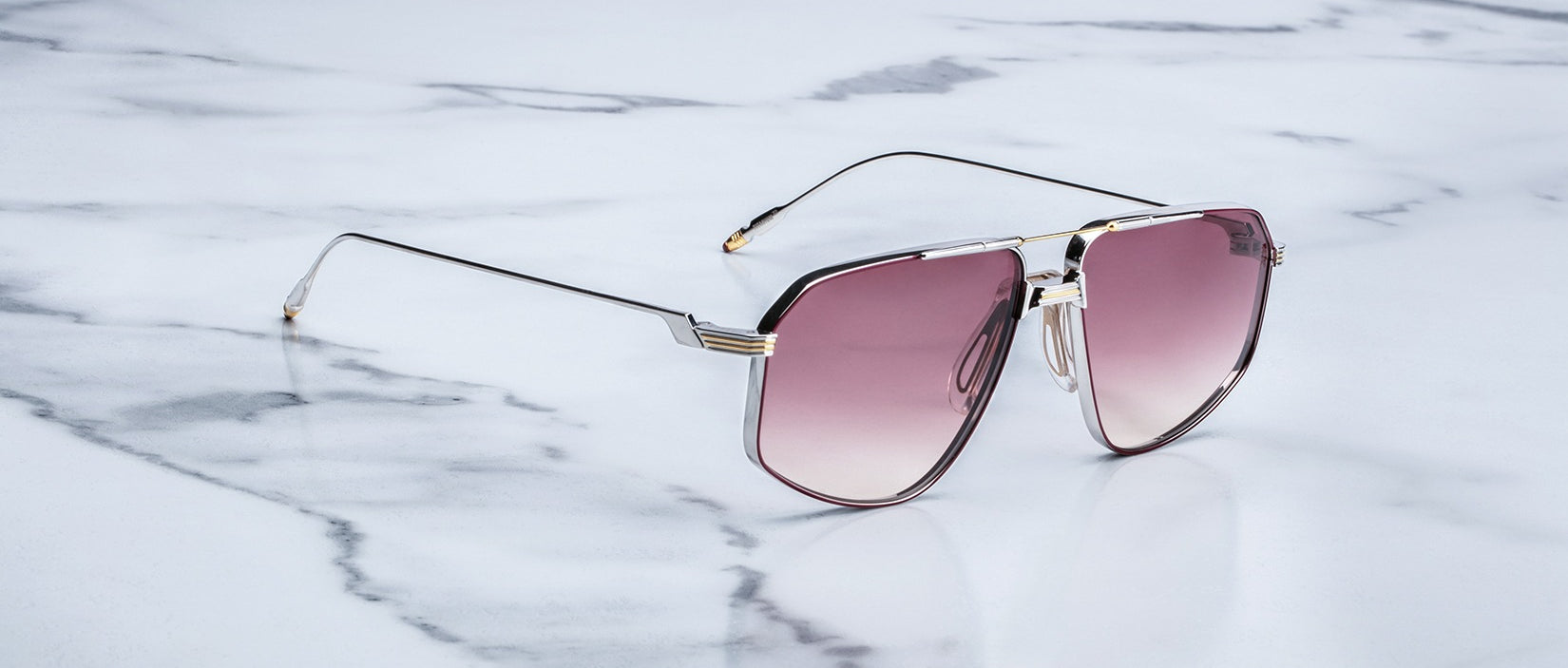 A pair of Jacques Marie Mage Jagger oversized aviator sunglasses by Jacques Marie Mage, featuring thin beta titanium frames and pink-tinted lenses, sits on a white marble surface.