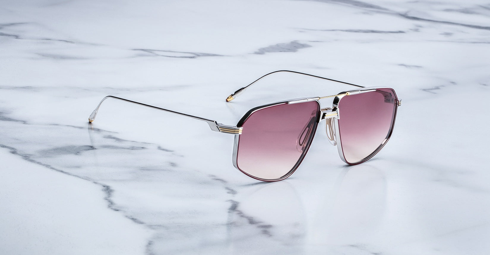 A pair of Jacques Marie Mage Jagger oversized aviator sunglasses by Jacques Marie Mage, featuring thin beta titanium frames and pink-tinted lenses, sits on a white marble surface.