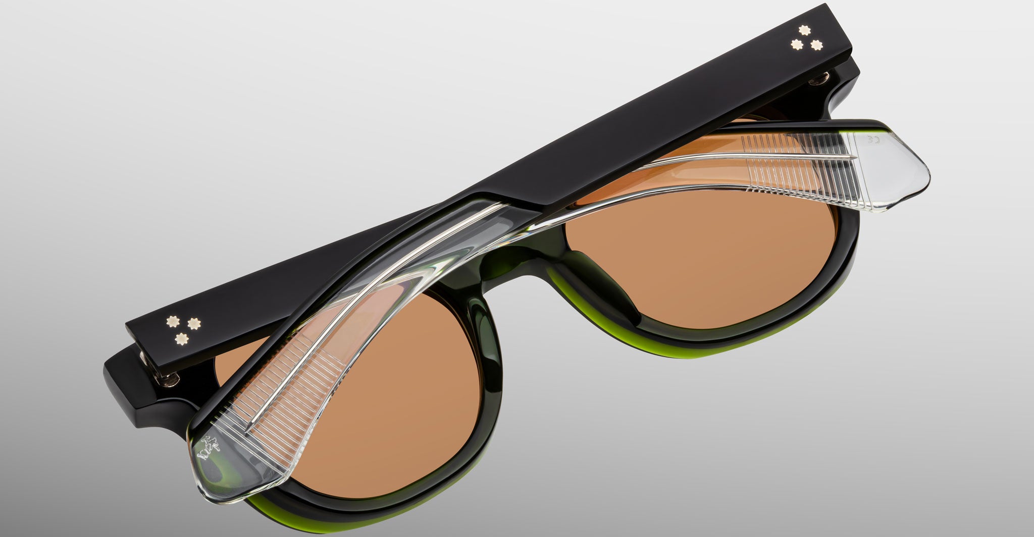 The Jacques Marie Mage Jax 2 sunglasses feature black, green, and clear plastic frames, brown-tinted lenses, and two small white floral designs on the temples, shown against a light gray background.