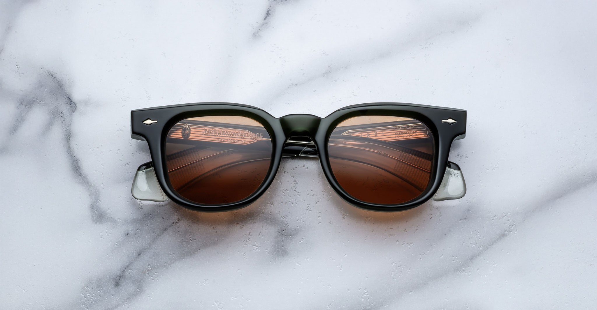 The Jacques Marie Mage Jax 2 sunglasses, featuring black frames and brown-tinted lenses, rest on a white marble surface. The lenses reflect patterns reminiscent of overhead lights or architectural details.