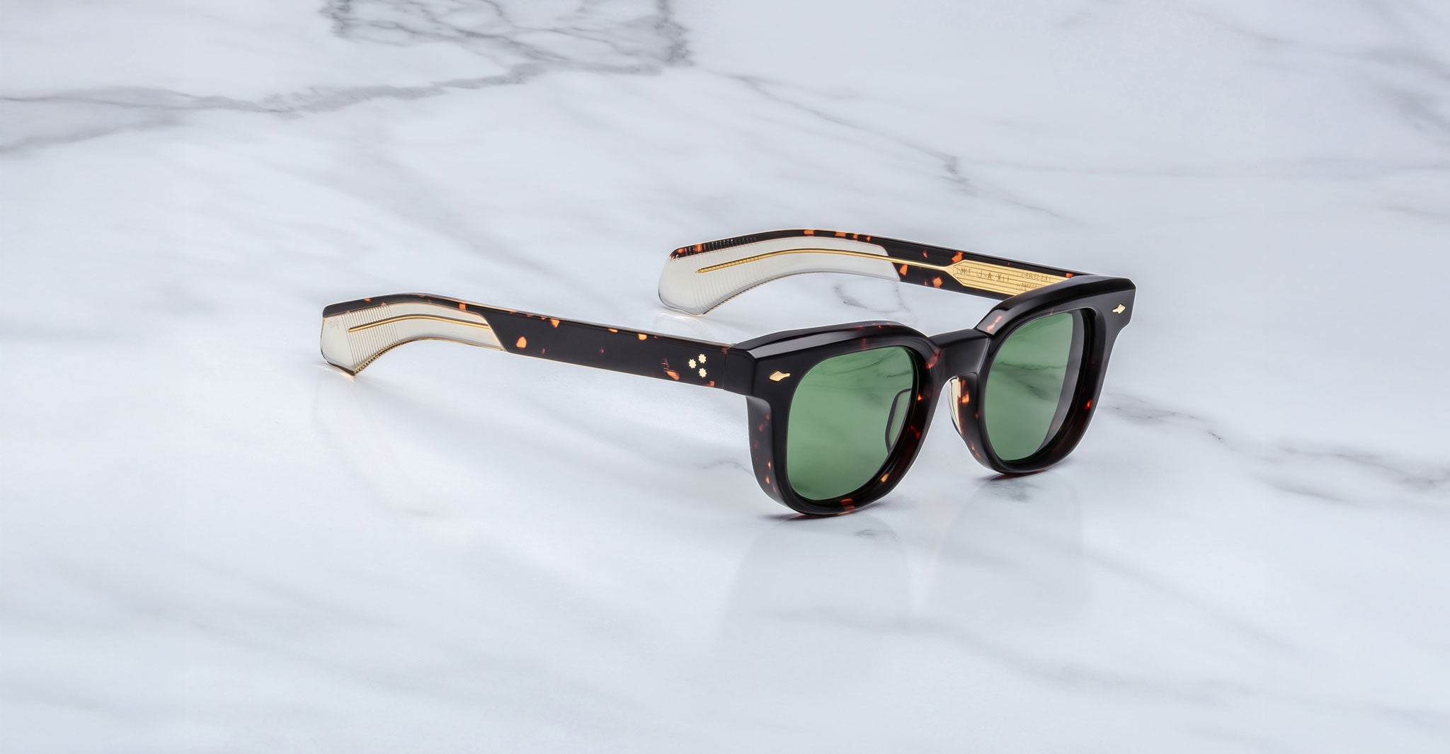 A pair of Jacques Marie Mage Jax 2 tortoiseshell sunglasses with green lenses and thick, straight arms rests on a white marble surface.