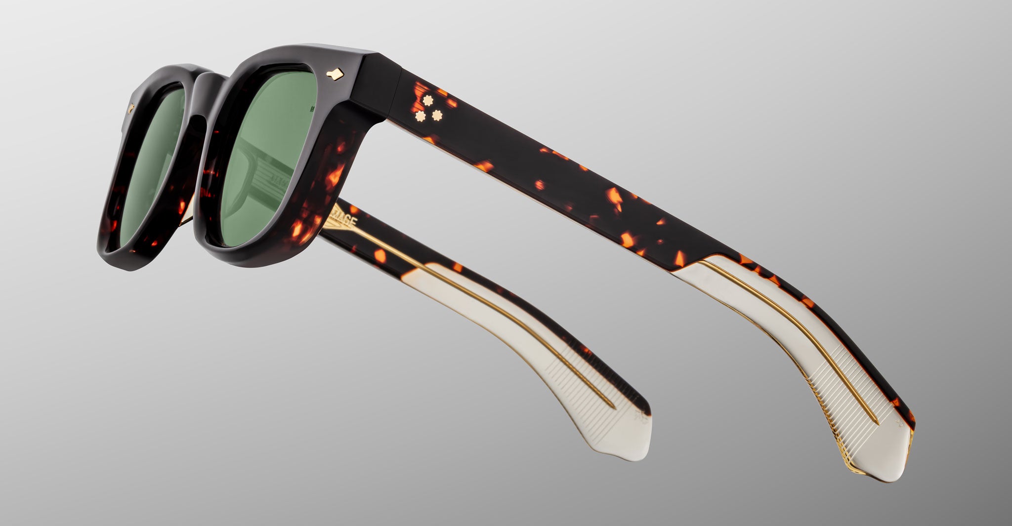 The Jacques Marie Mage Jax 2 sunglasses feature thick, dark tortoiseshell frames, green-tinted lenses, and temples with gold detailing and a textured inner section, displayed against a light gray background.