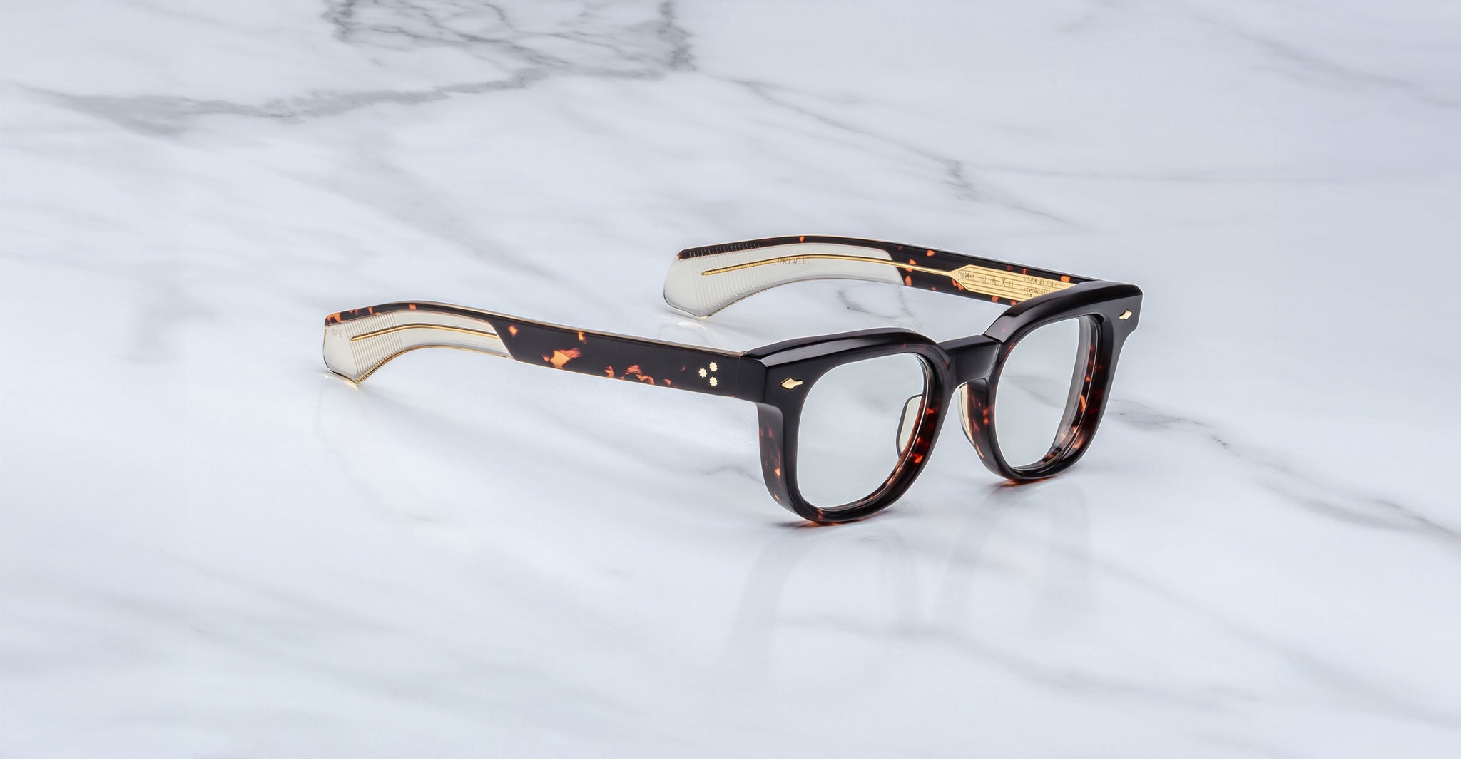 The Jacques Marie Mage Jax 2 eyeglasses feature thick-rimmed, rectangular frames in a tortoiseshell pattern with gold-accented temples, displayed on a white marble surface.