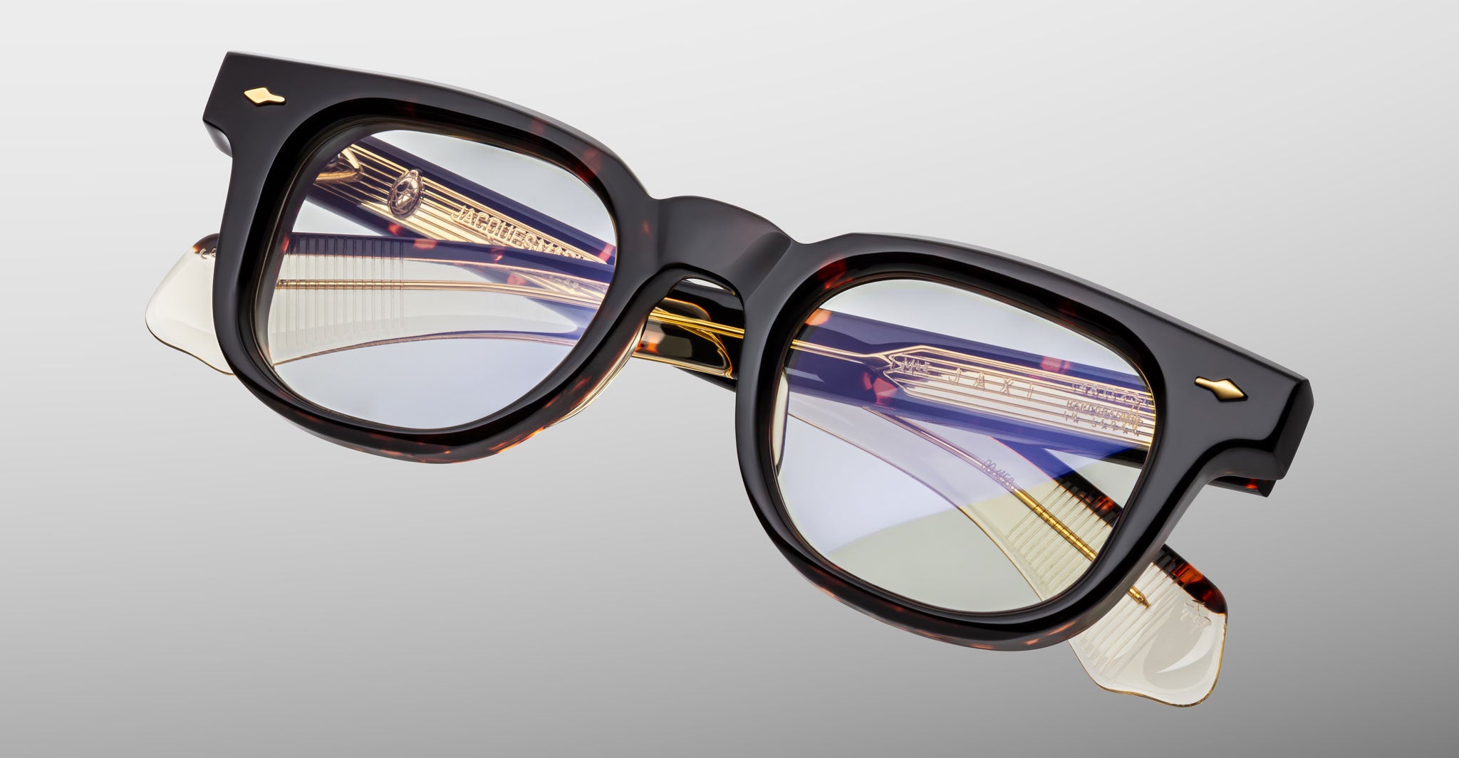 The Jacques Marie Mage Jax 2 eyeglasses feature thick-rimmed, rectangular black and tortoiseshell frames with gold accents and clear lenses, shown against a light gray background.