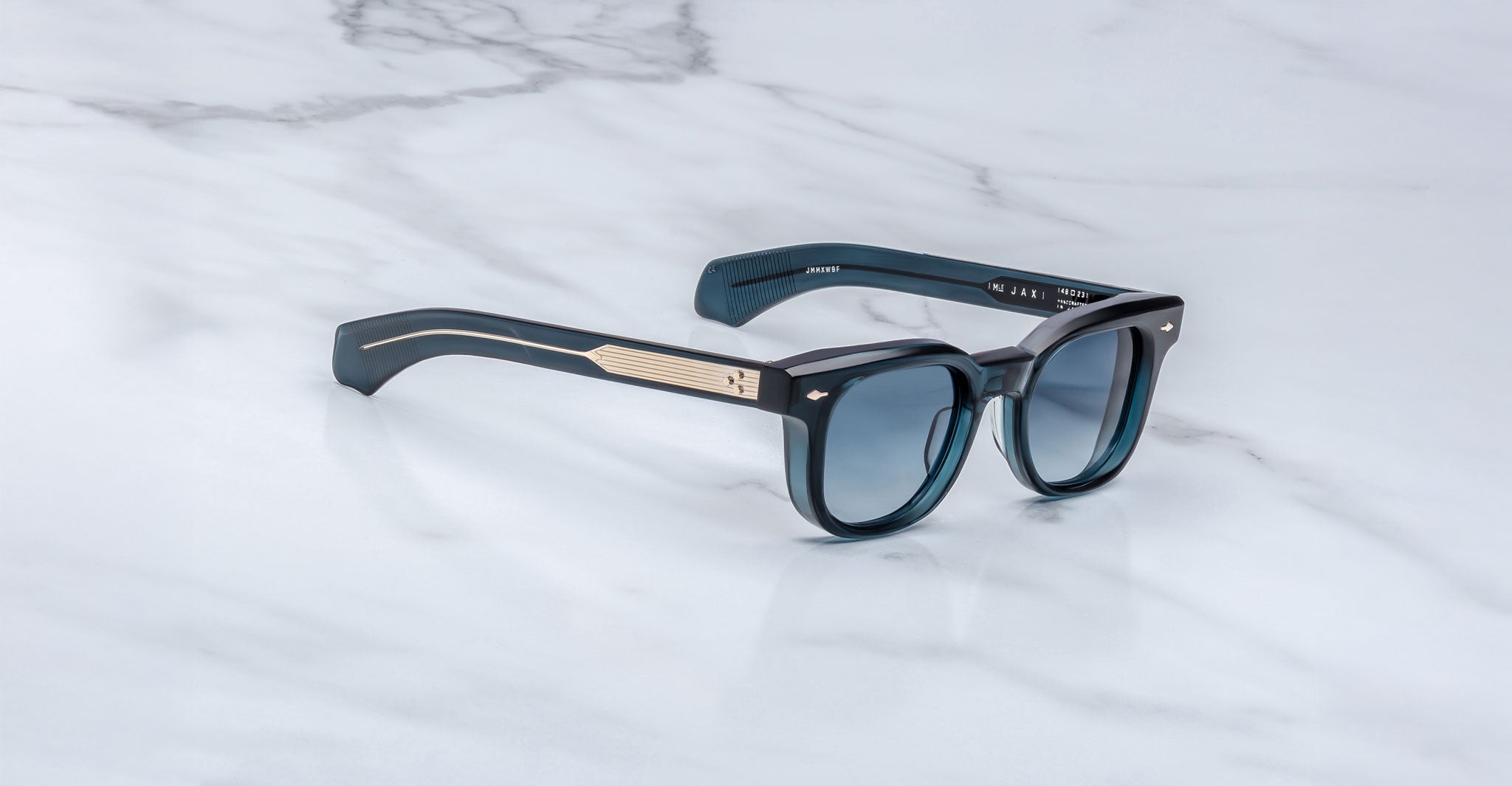 The Jacques Marie Mage Jax 2 is a pair of black rectangular sunglasses with blue-tinted lenses and gold accents on the arms, displayed on a white marble surface.