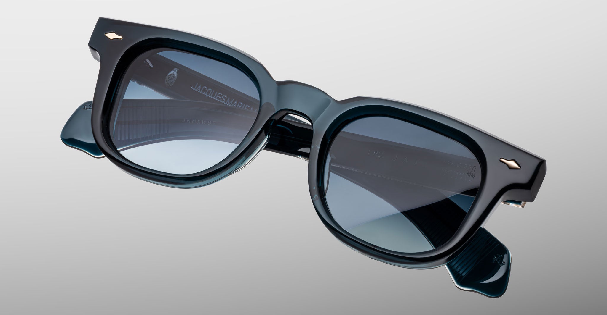 The Jacques Marie Mage Jax 2 sunglasses feature dark blue rectangular frames with dark tinted lenses and metallic arrow-shaped accents at the corners, displayed against a light grey background.