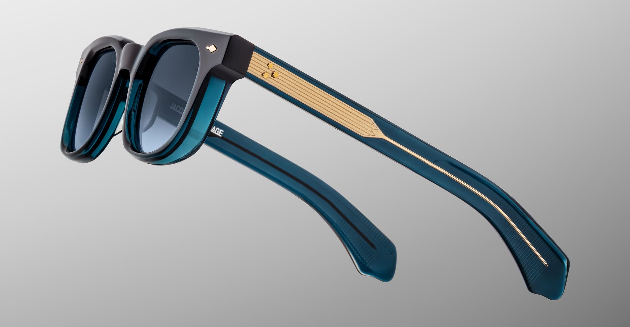 The Jacques Marie Mage Jax 2 sunglasses feature thick, dark blue translucent frames, gold temple accents, and dark tinted lenses, shown against a plain grey background.
