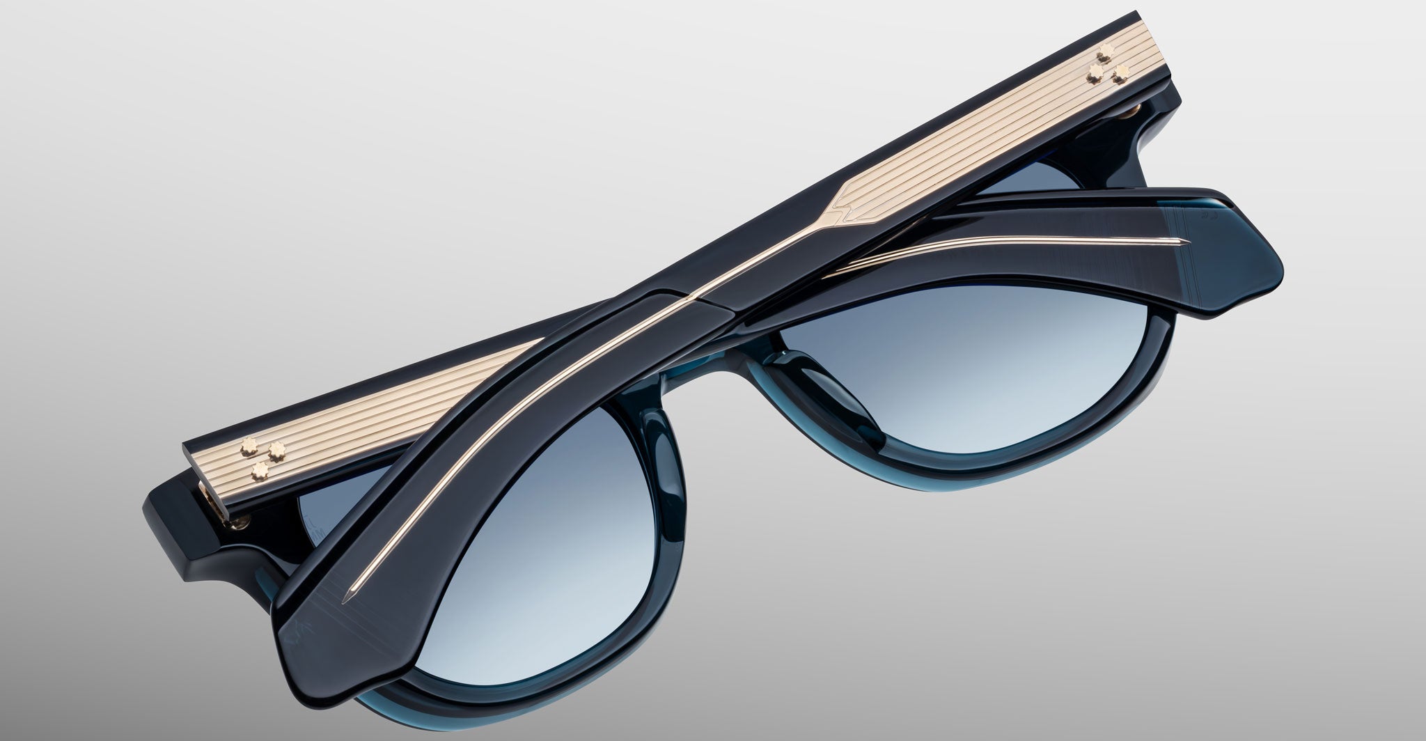 The Jacques Marie Mage Jax 2 sunglasses feature dark blue frames, blue-tinted lenses, and gold metal detailing along the temples, shown folded from above on a gradient gray background.