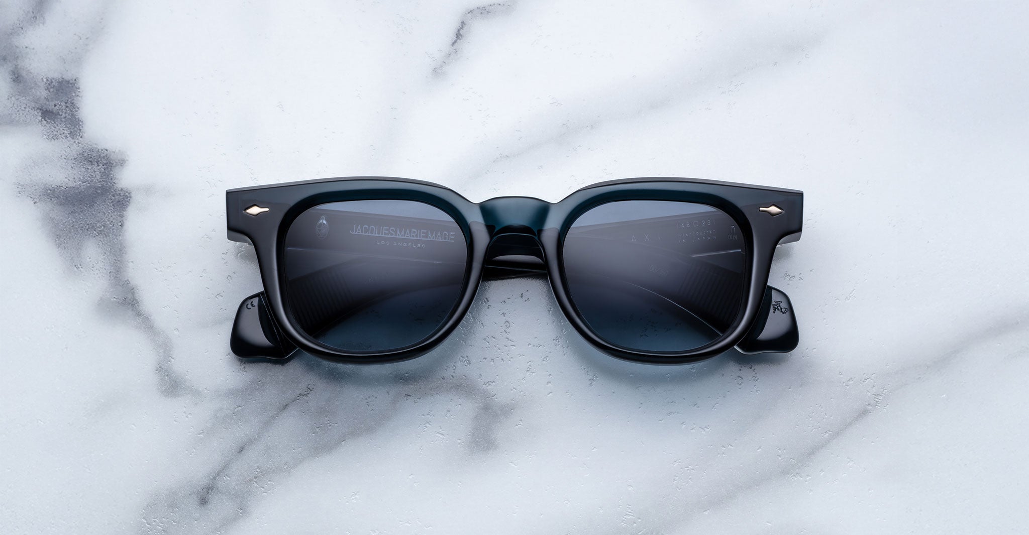 The Jacques Marie Mage Jax 2 black square-shaped sunglasses with dark lenses are placed on a white marble surface with gray veining, facing up to display their front frames and lenses.