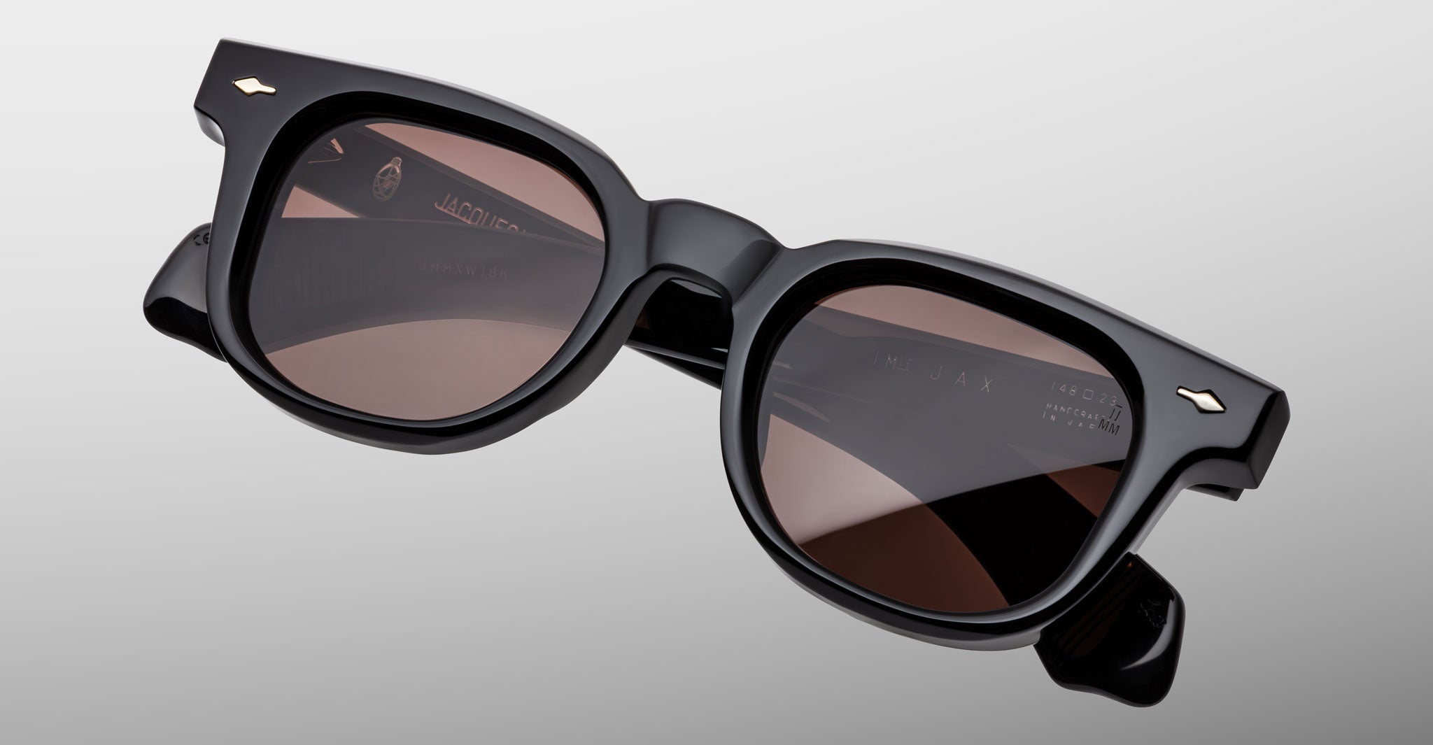 A pair of Jacques Marie Mage Jax 2 black, thick-rimmed sunglasses with slightly rounded square lenses and dark brown tint, displayed on a plain light gray background.