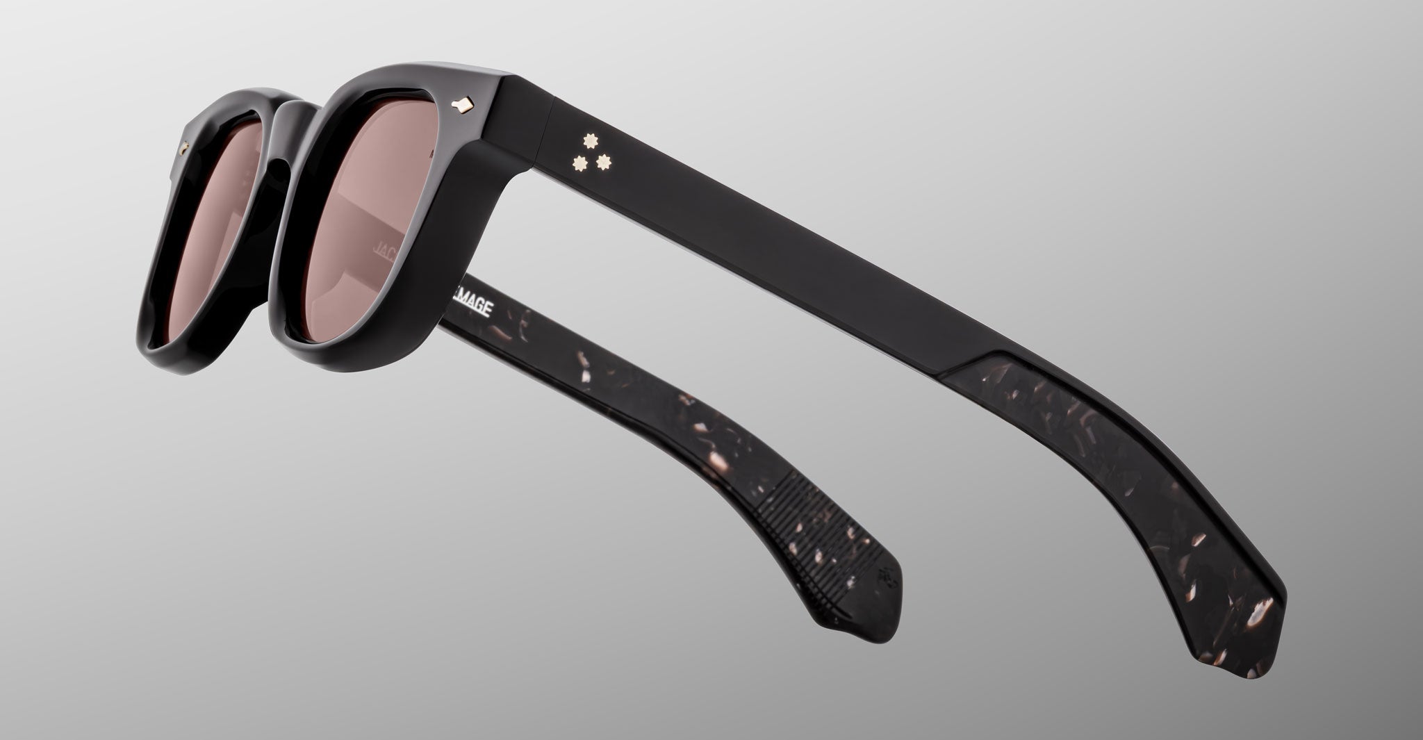 The Jacques Marie Mage Jax 2 sunglasses in black, featuring dark lenses and small flower-shaped accents on the upper corners, are shown folded with one arm across the front on a light gray background.