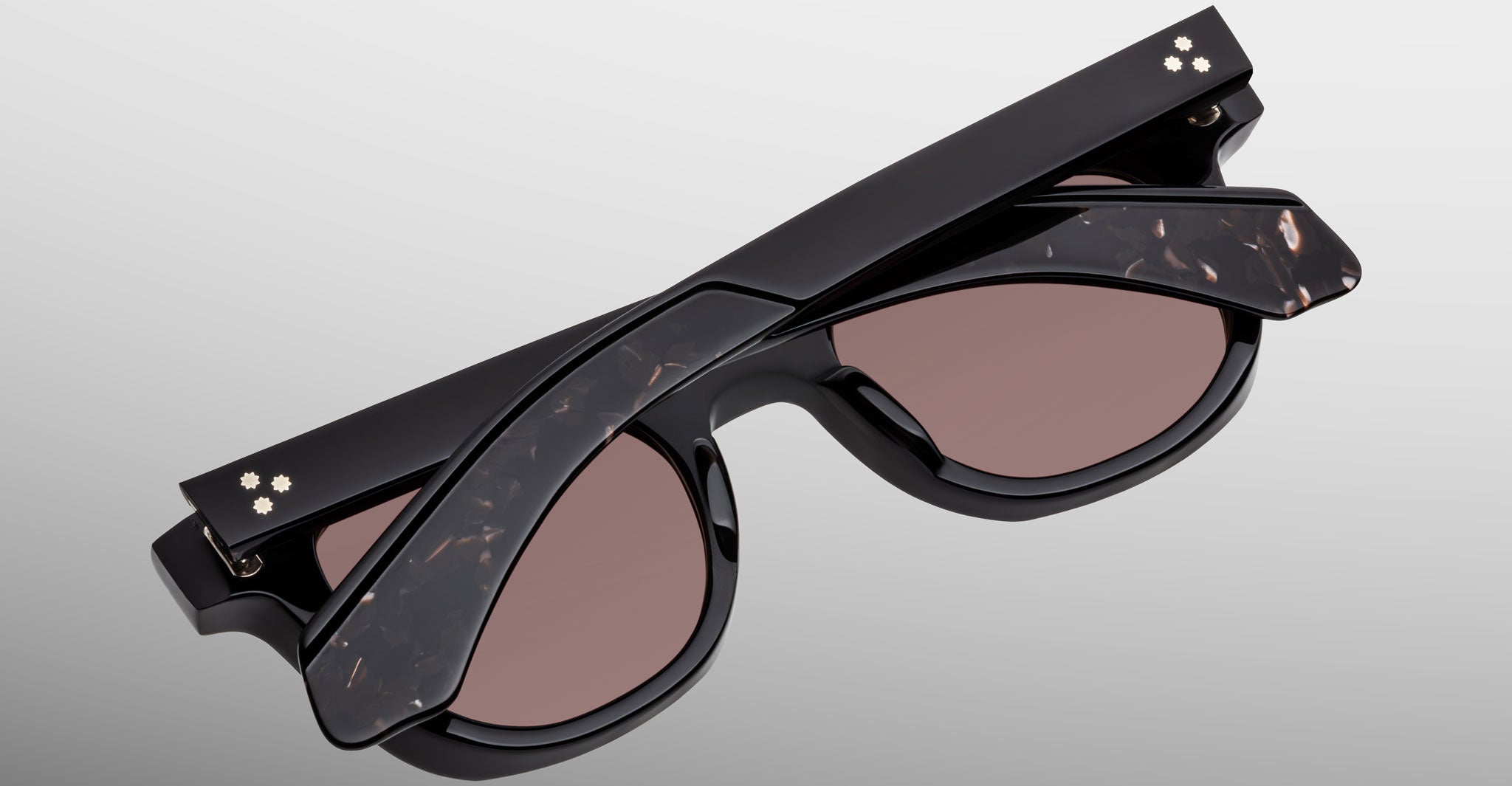 The Jacques Marie Mage Jax 2 sunglasses are shown folded, featuring a black frame with marbled arm accents and two small flower-shaped gold details at the temple corners. The lenses are brown.