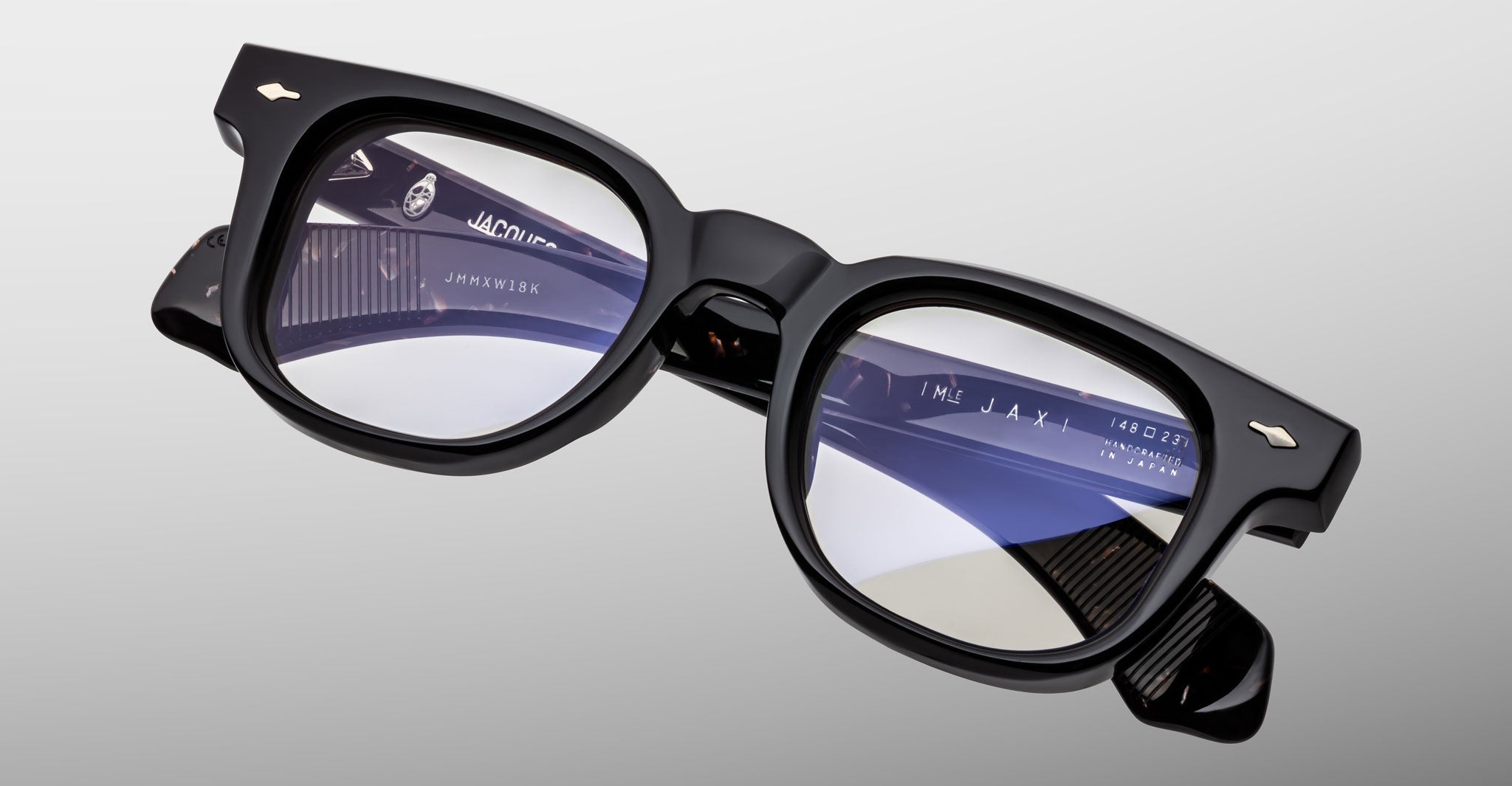 The Jacques Marie Mage Jax 2 eyeglasses, featuring black thick rims and rectangular lenses, are displayed on a light gray background. Blue and purple reflections appear on the left lens, with product details inside the arms visible.