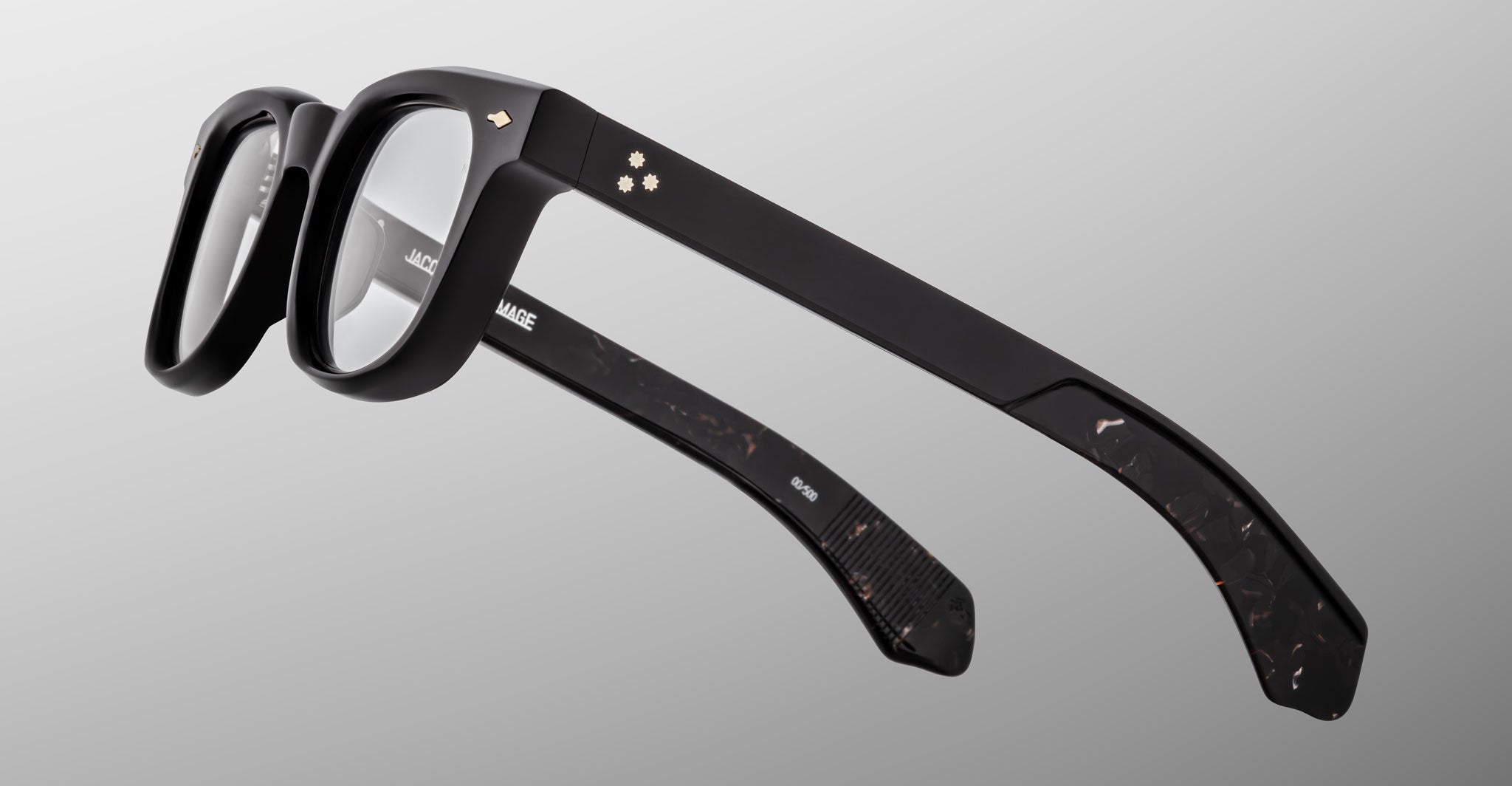 The Jacques Marie Mage Jax 2 eyeglasses feature black thick frames and clear lenses, shown from a rear side angle on a plain light gray background. Subtle decorative details appear near the hinges on the arms.