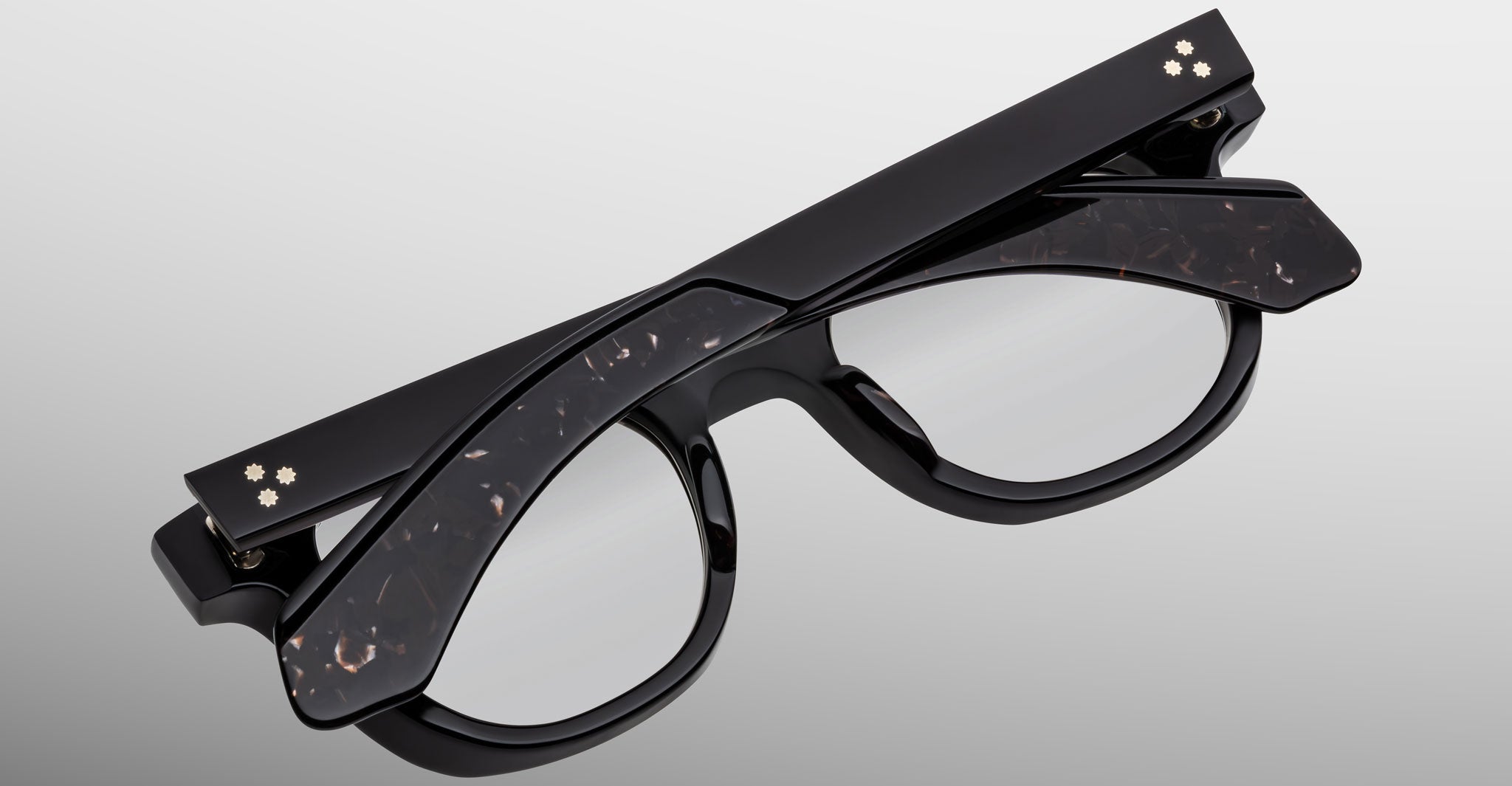 The Jacques Marie Mage Jax 2 eyeglasses, featuring black rectangular frames, are folded with the temples crossed over the lenses and placed on a light gray background.